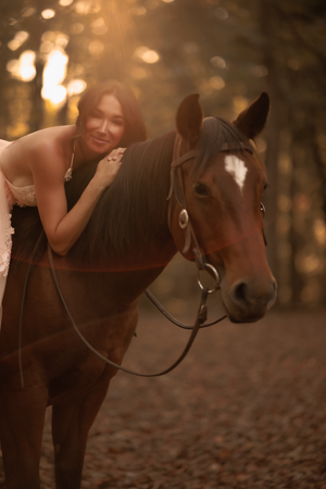 A brown horse with a white blaze stands in a sun-dappled forest while being gently embraced by its rider.