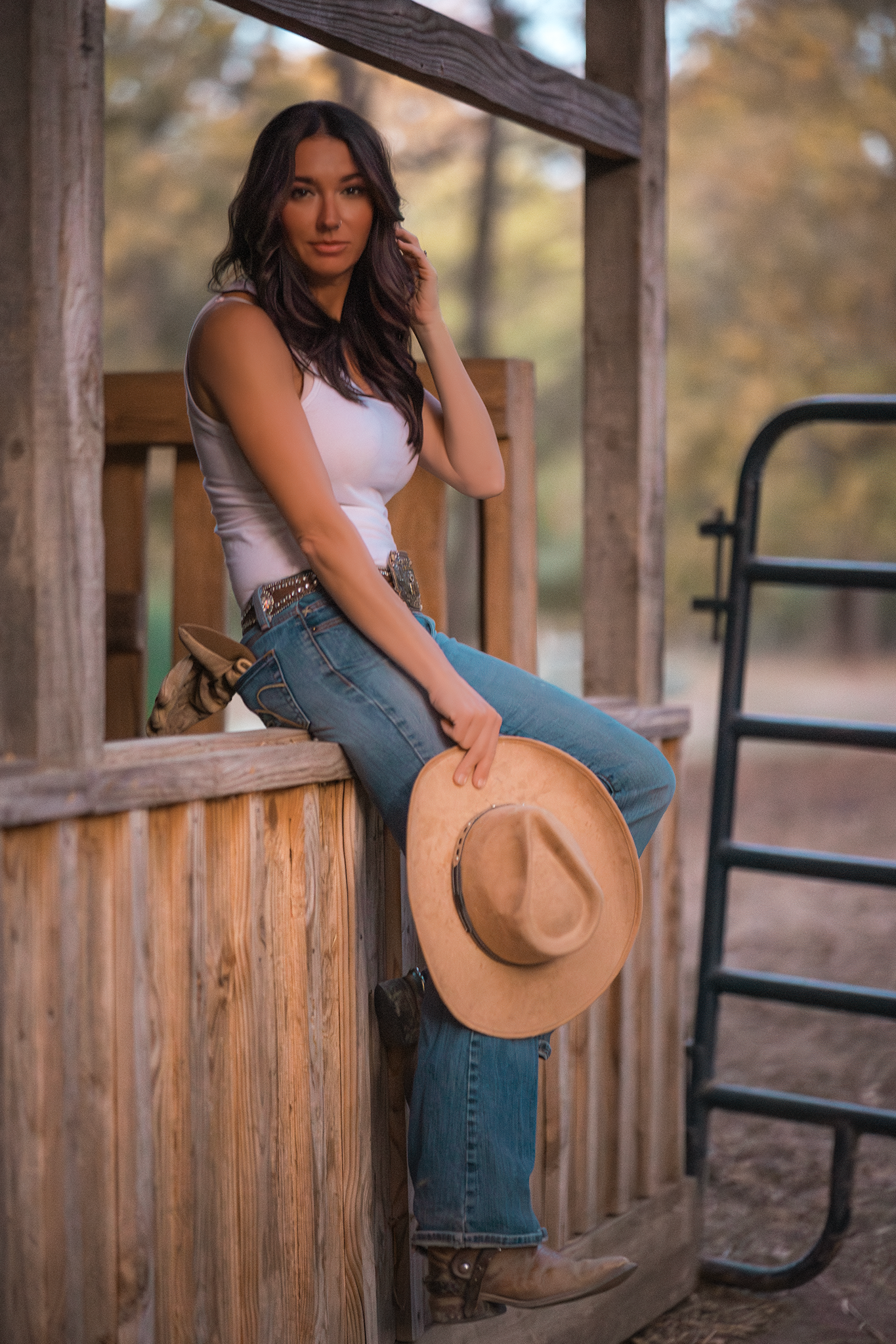 A woman in western attire with jeans and white tank top sits on a wooden railing holding a tan cowboy hat at sunset.