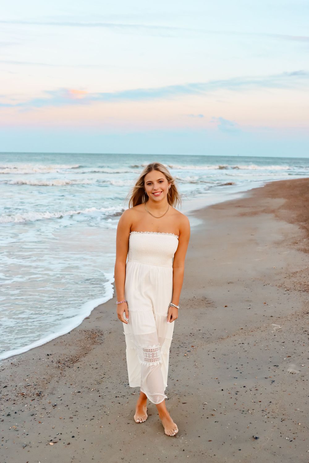 Woman in white dress walks along beach shoreline during sunset.