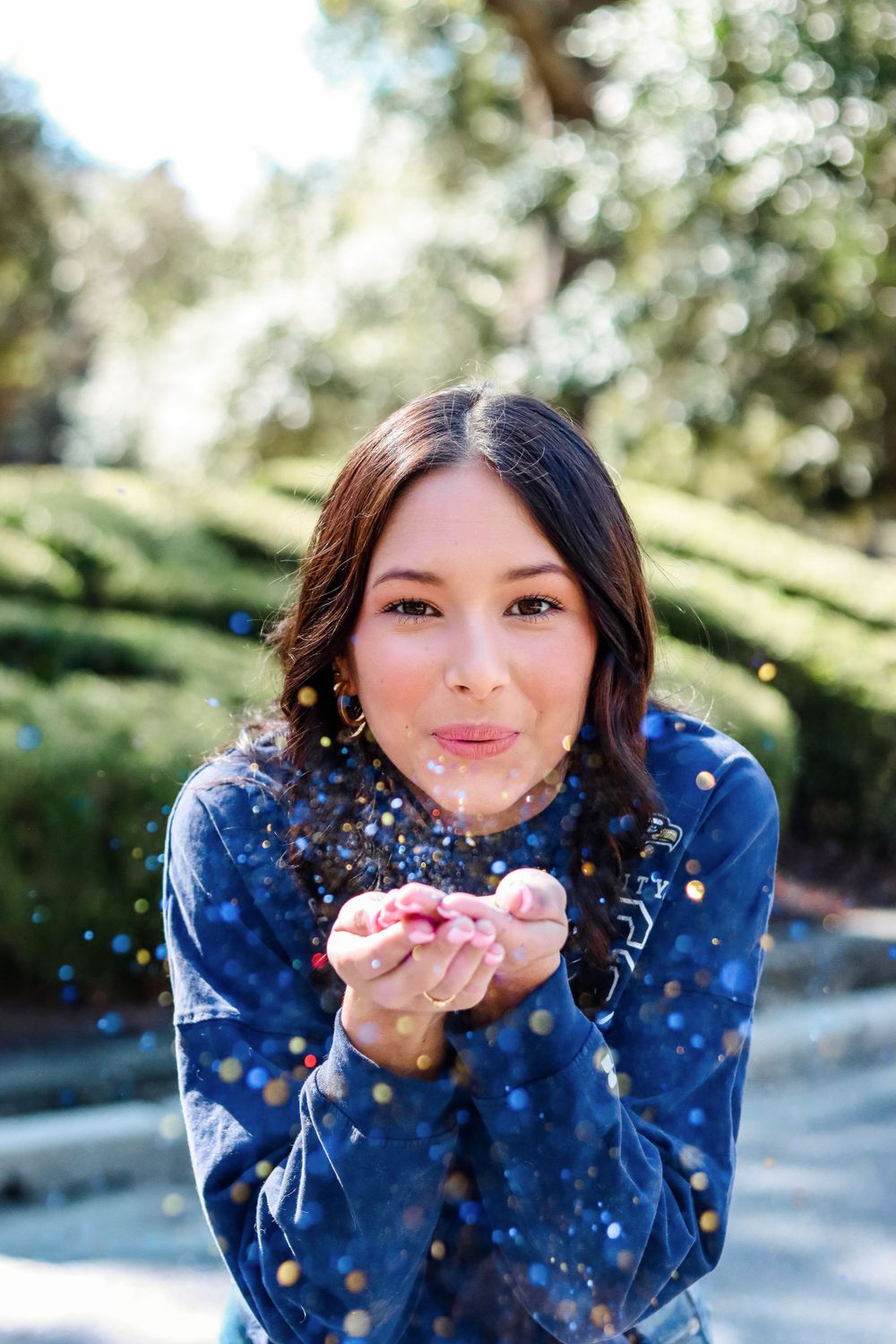 Woman in blue sweater blows colorful glitter confetti from her hands in outdoor portrait.