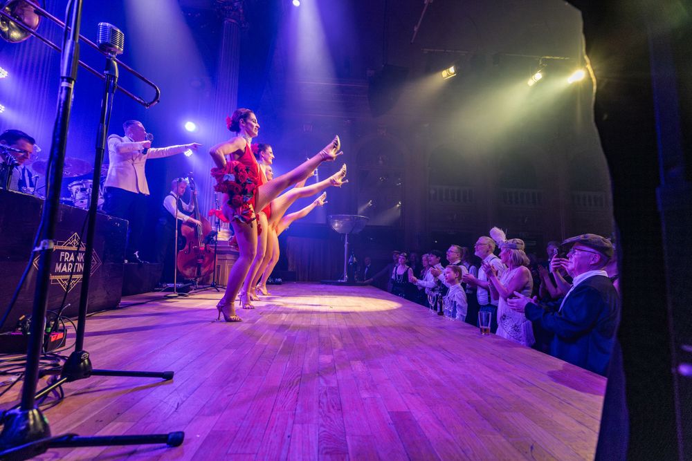 Musicians perform on stage under dramatic purple lighting while an audience watches from the side at an intimate concert venue.