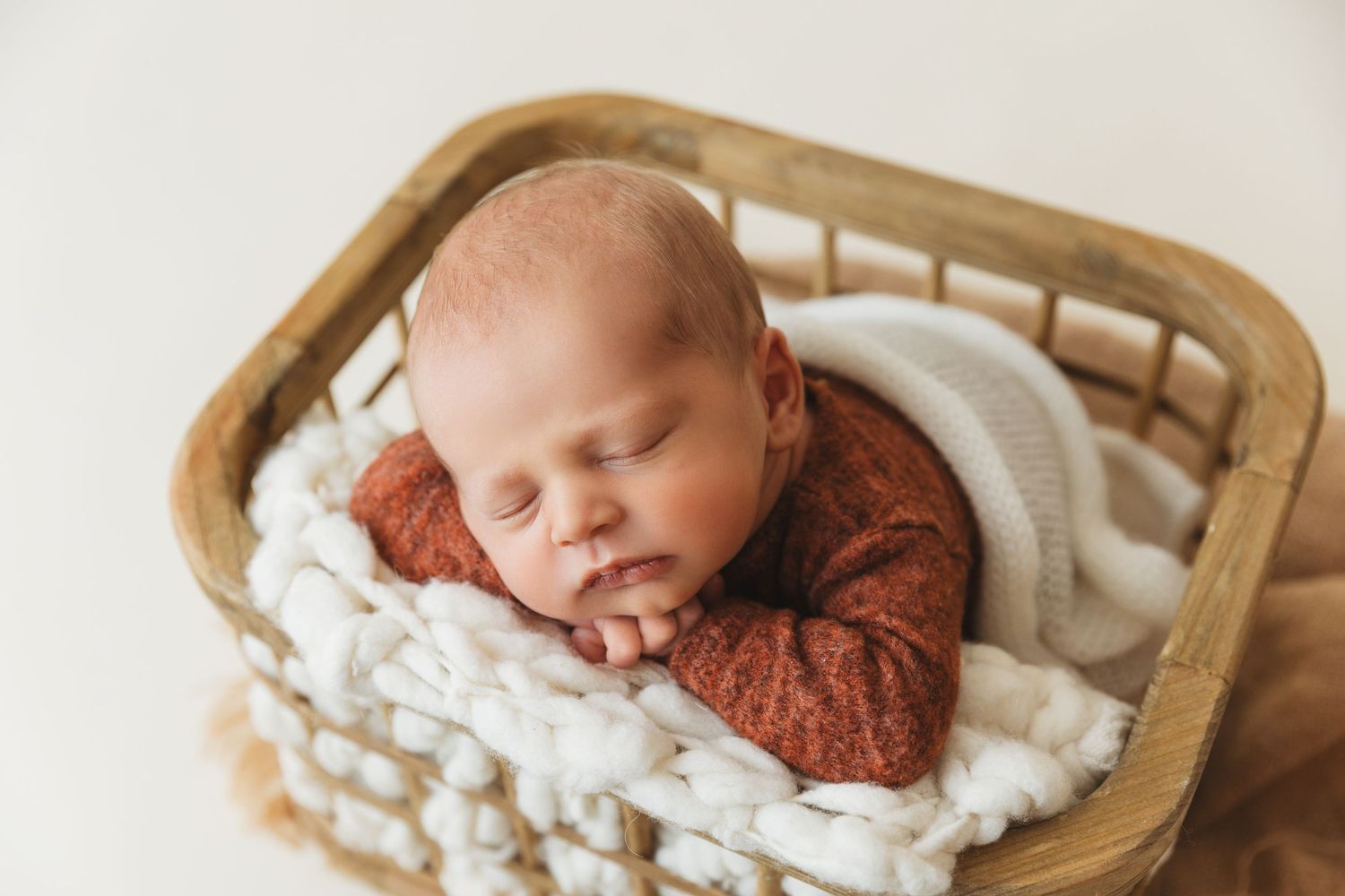 Sleeping newborn in rust-colored outfit nestled in wooden basket with white padding.