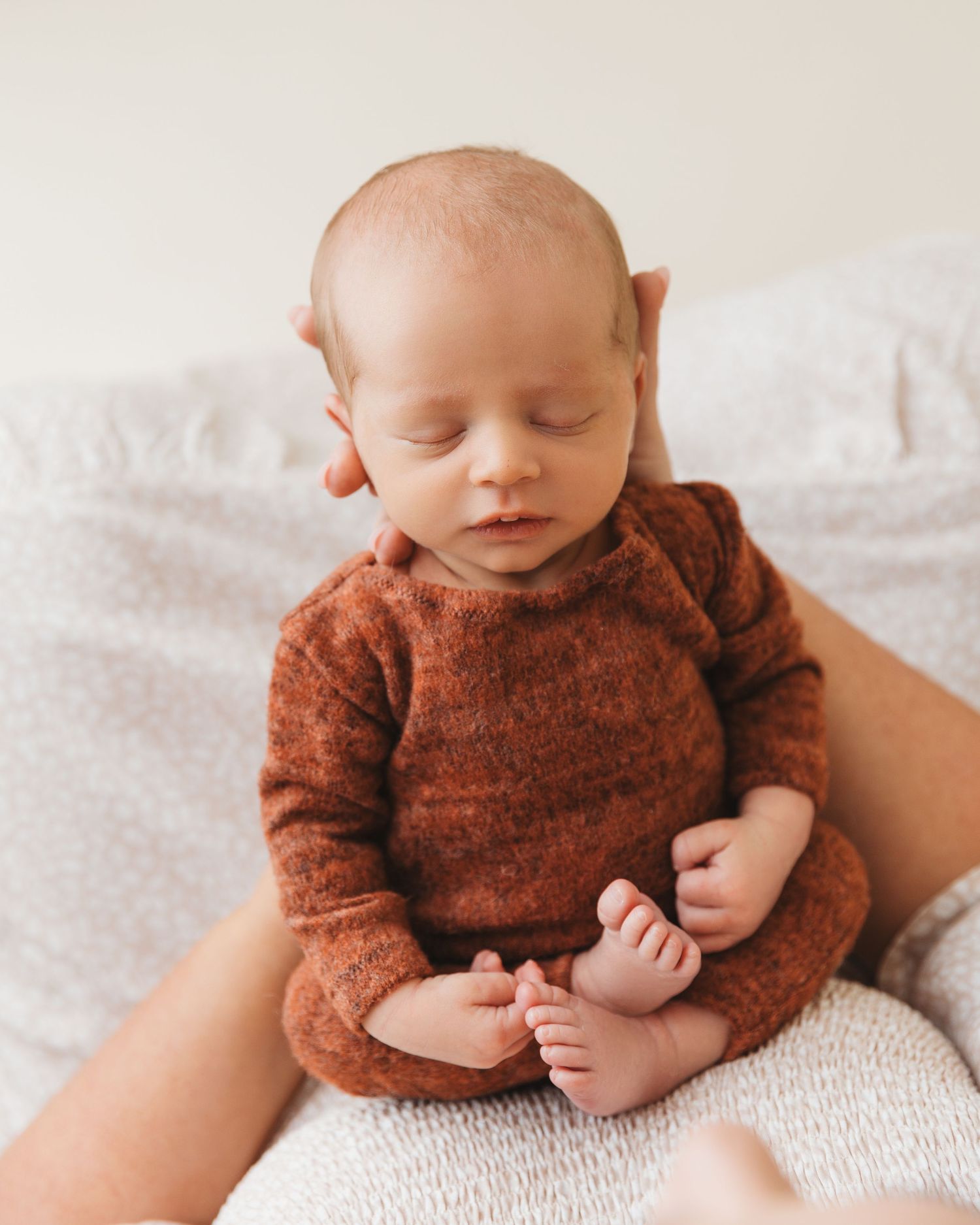 Peaceful newborn wearing brown knit sweater resting on white bedding.