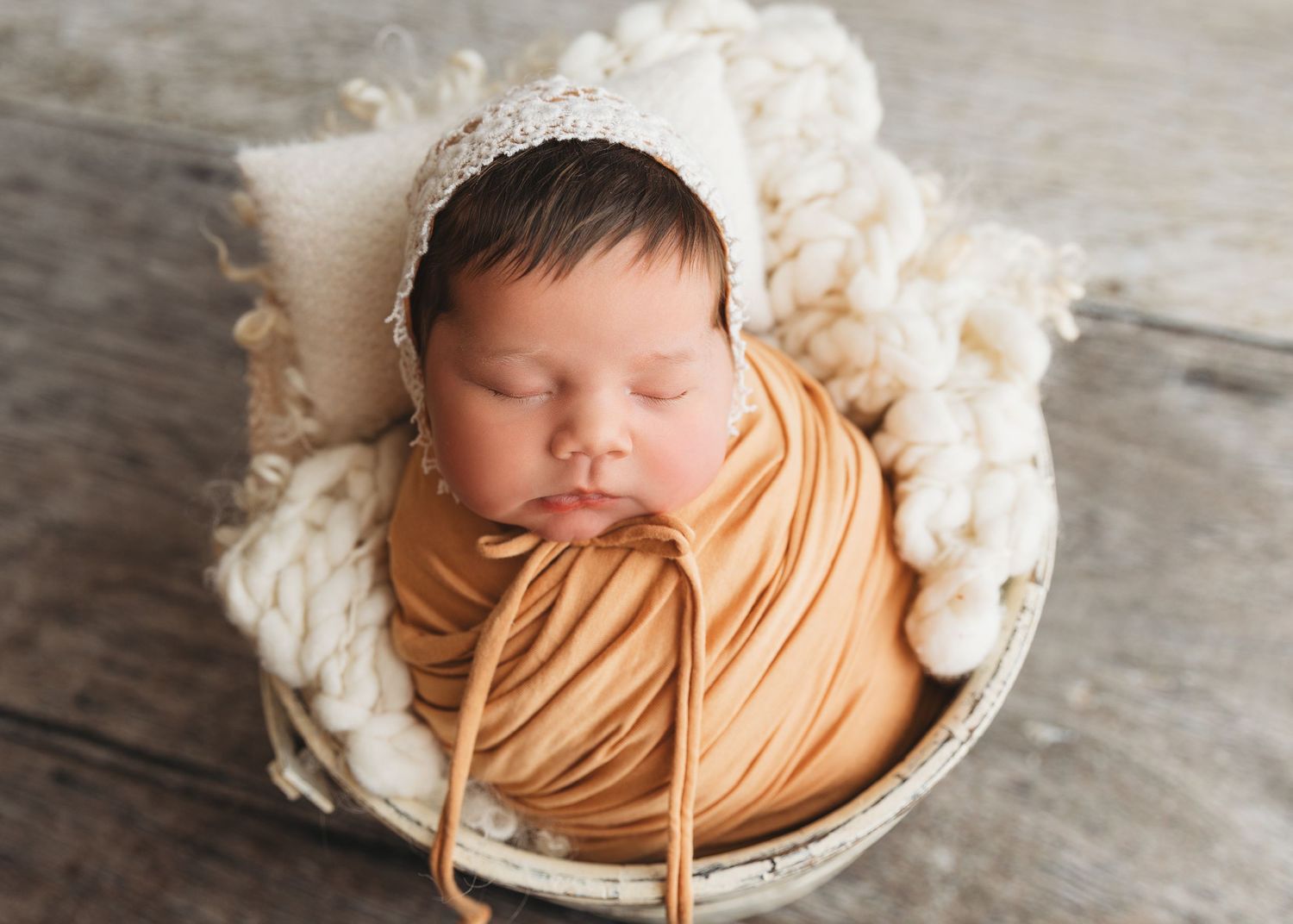 Sleeping newborn wrapped in caramel-colored fabric in white basket against wooden backdrop.