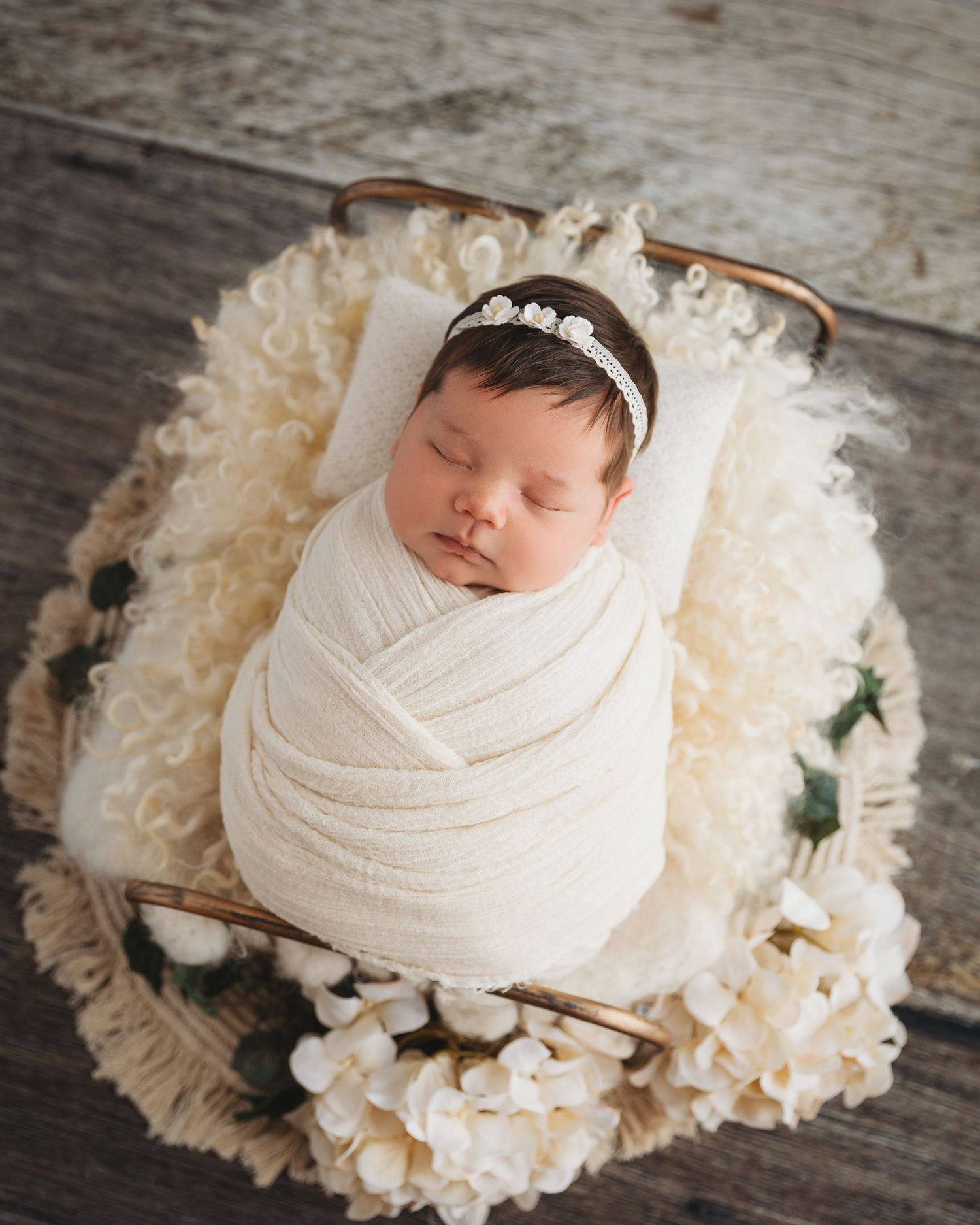 Newborn baby wrapped in white swaddle lying on cream floral arrangement against wooden backdrop.