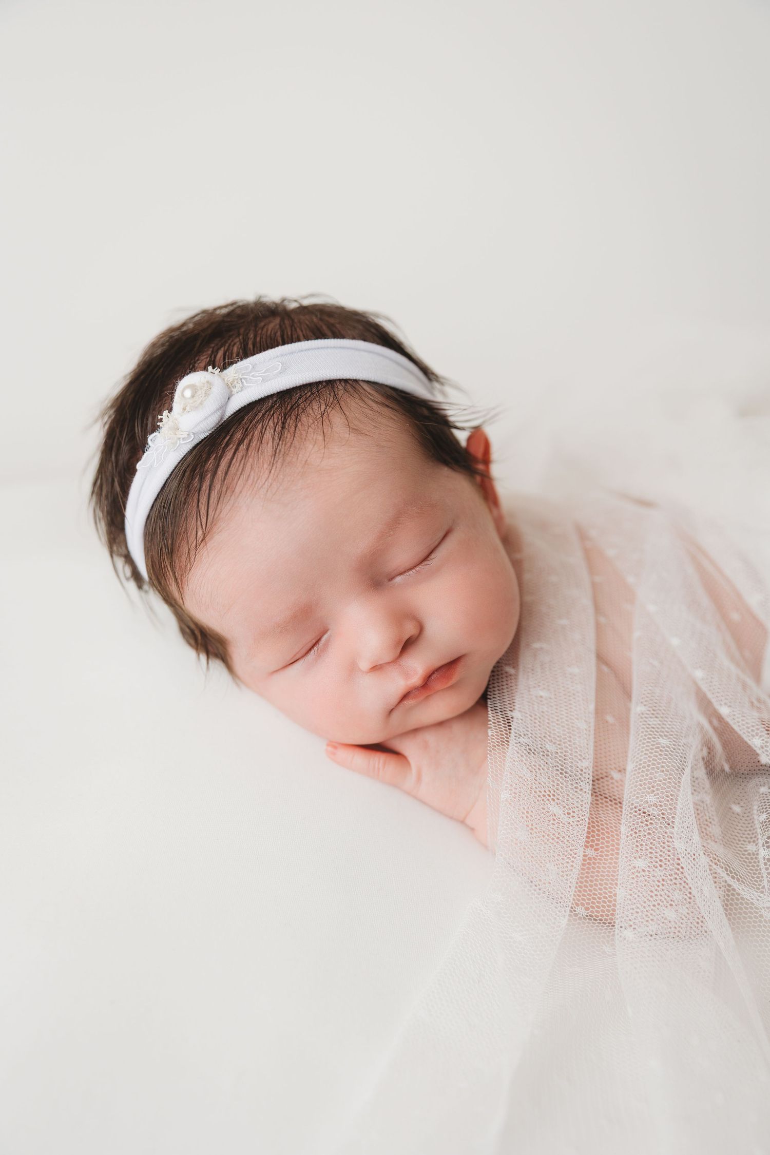 Sleeping newborn wearing delicate white lace wrap and headband against light background.