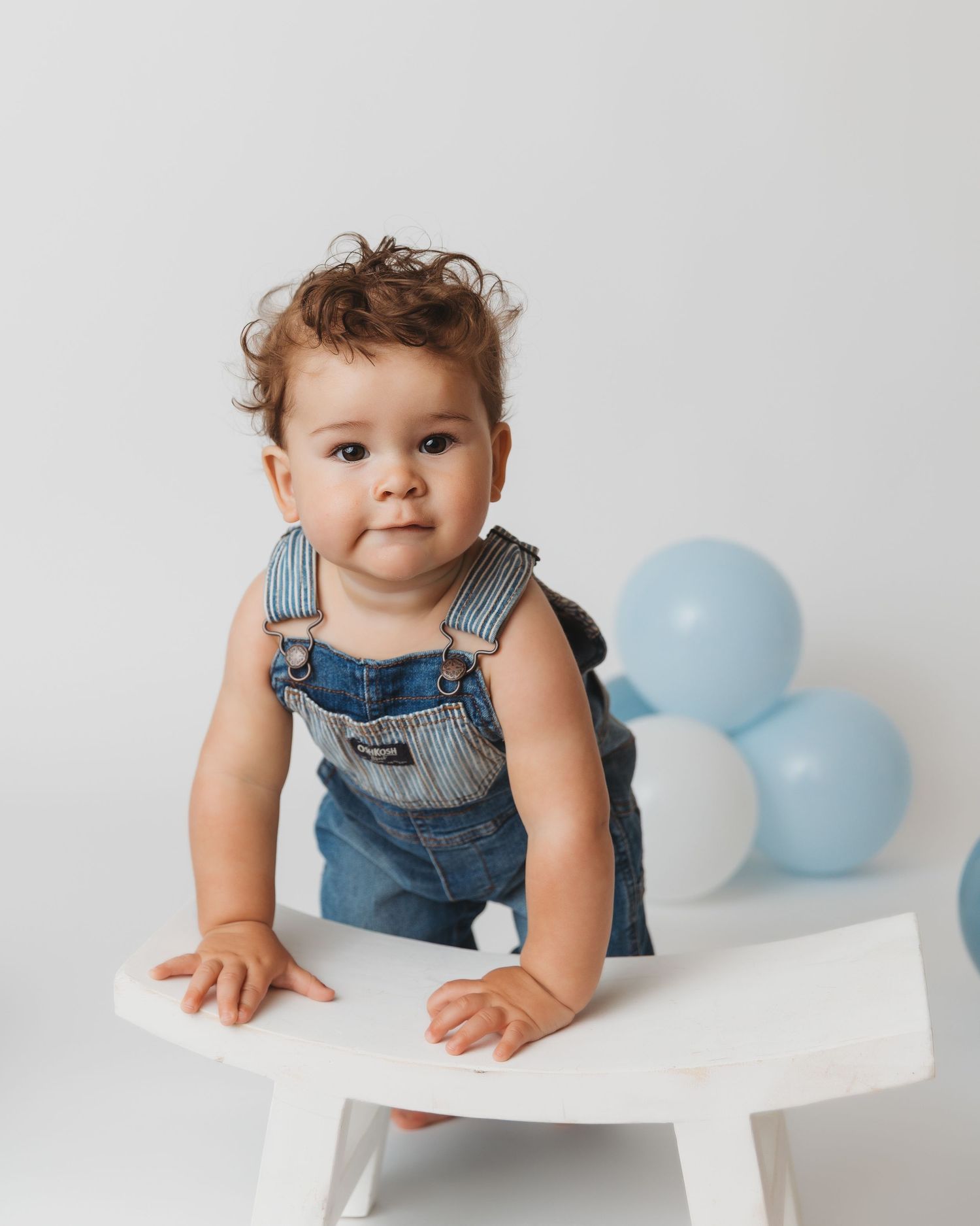Baby in striped overalls crawling on white table with blue balloons in background.