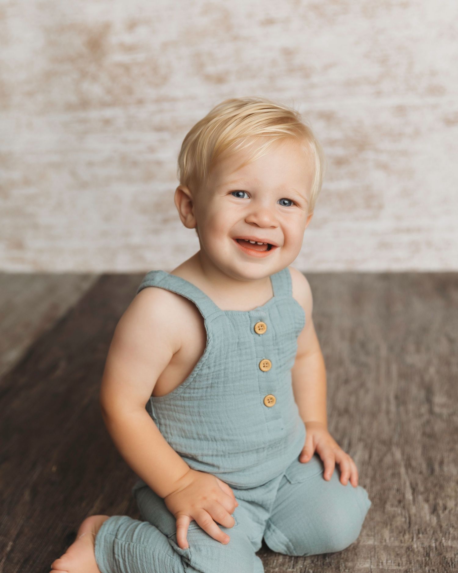 Baby wearing blue linen romper with wooden buttons on textured wood floor.