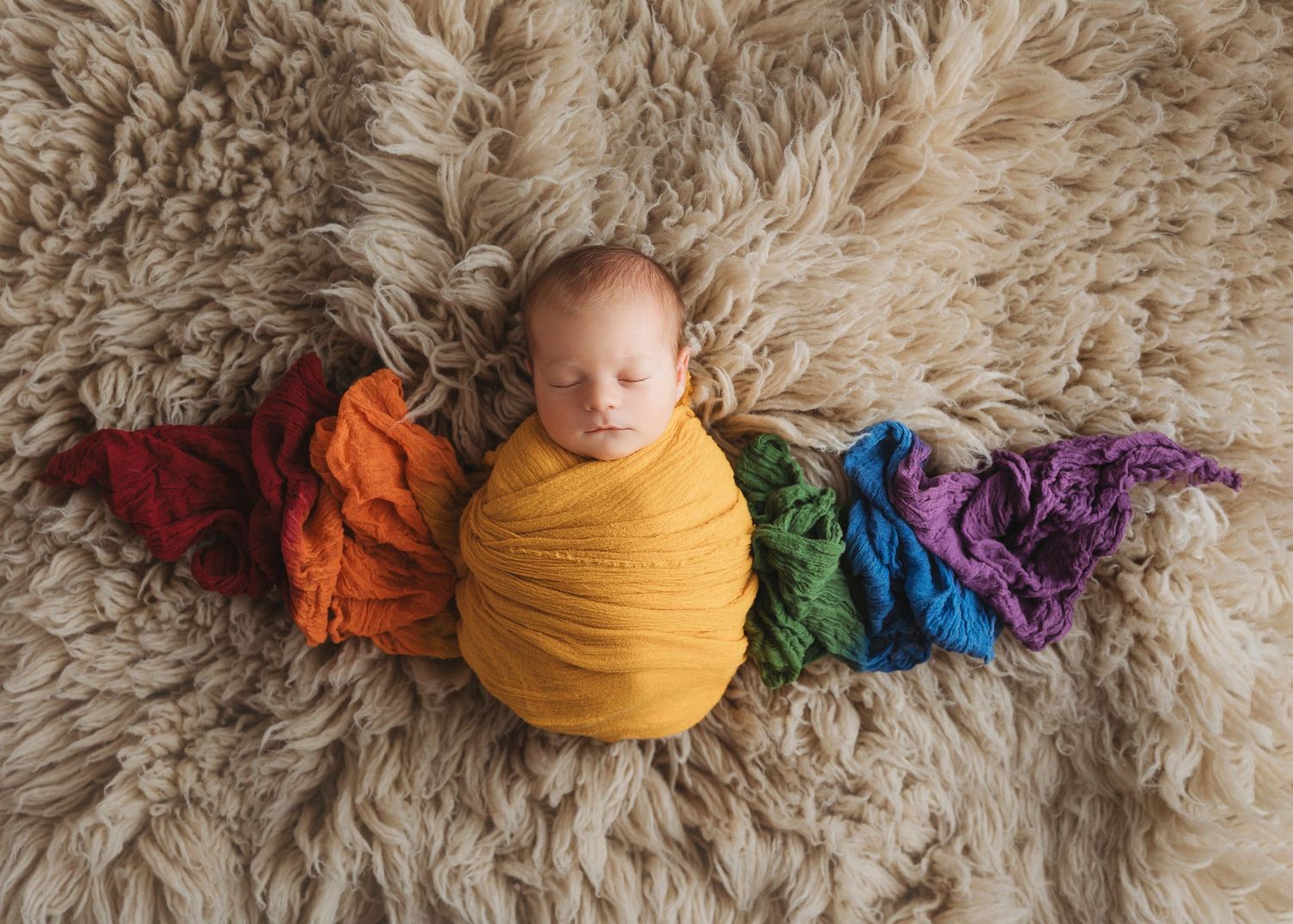 Newborn photography session with colorful rainbow wraps and textured beige fur backdrop.