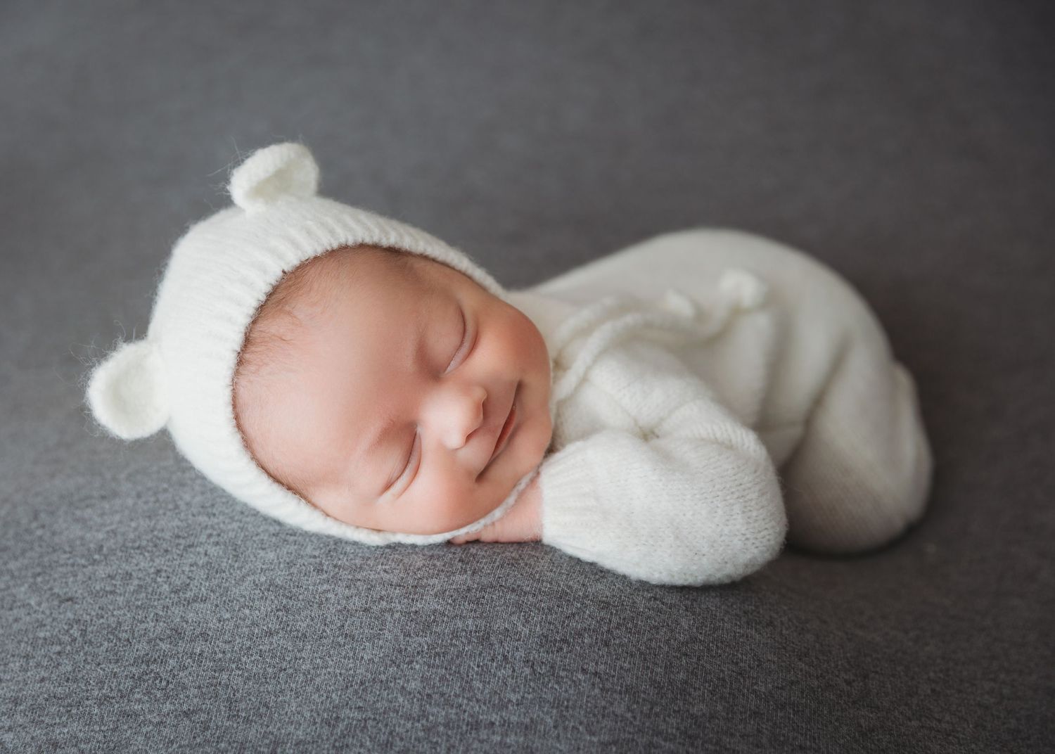 Peaceful newborn wearing white knit cap resting on gray textured background.