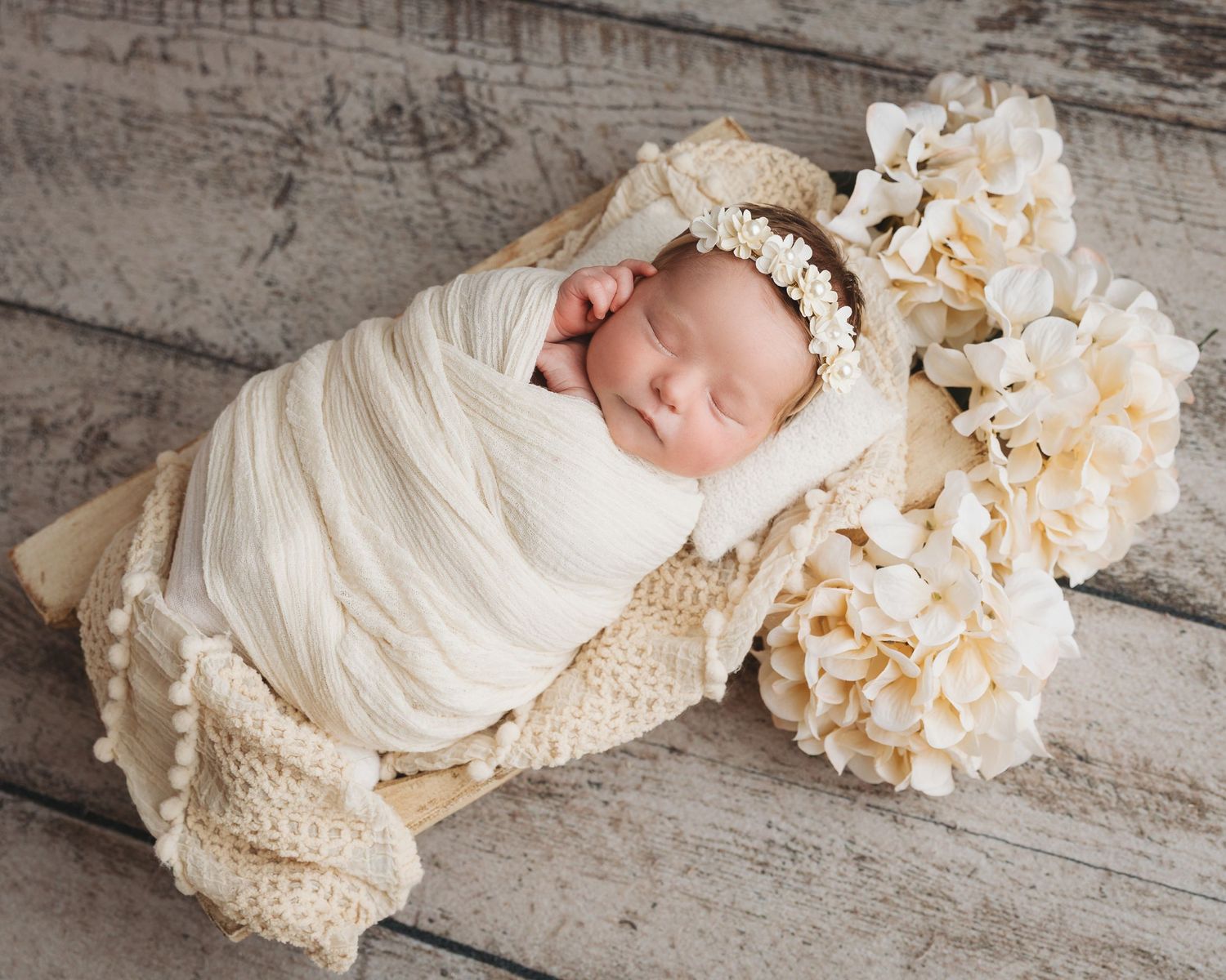 Newborn baby wrapped in cream fabric lying among soft floral arrangements on wooden surface.