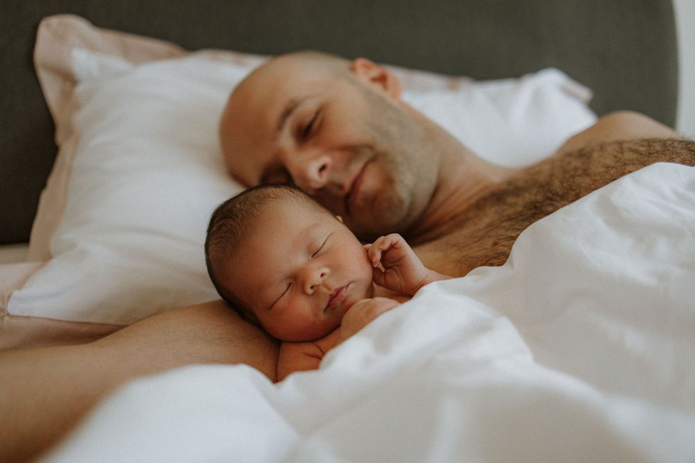 A baby and parent sleeping peacefully together in a white bed, with soft warm lighting.