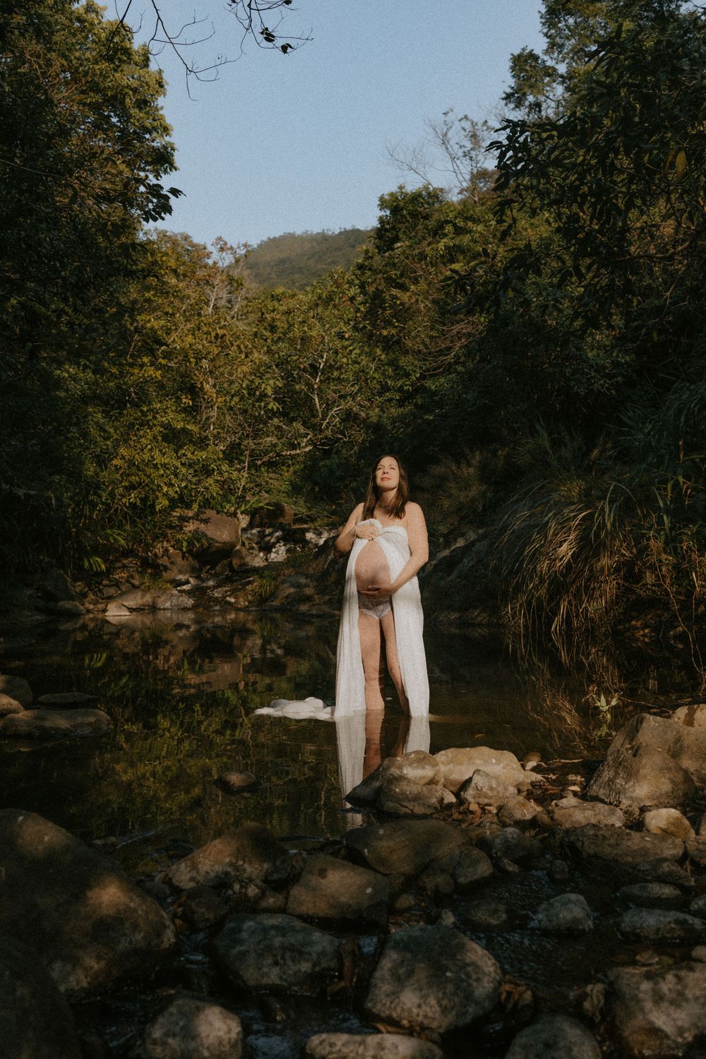 A figure in a flowing white dress stands on rocky riverbank at sunset surrounded by lush forest scenery.