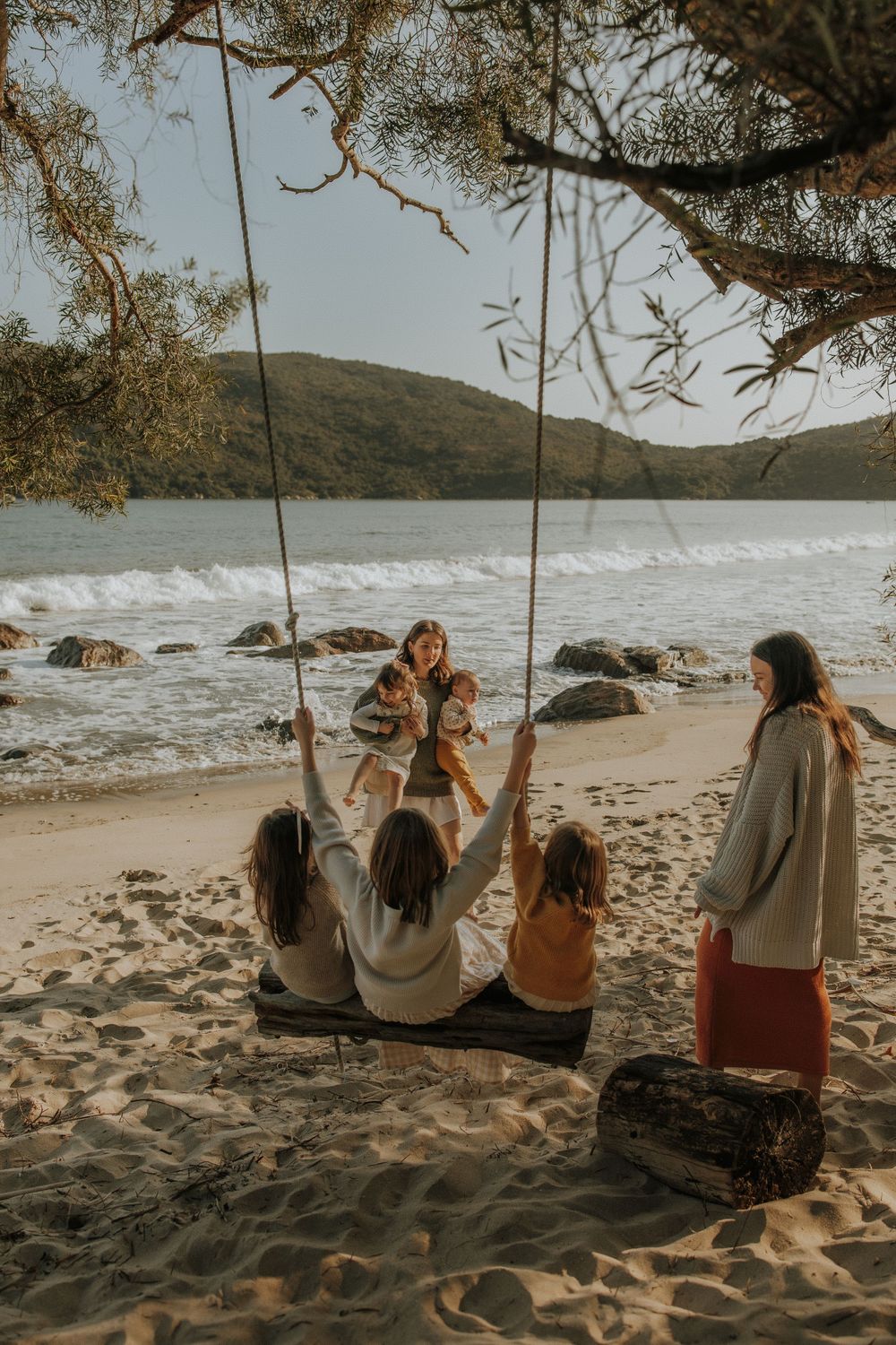 Group enjoying a beach swing on sandy shore with crashing waves and rolling hills in the background at sunset.