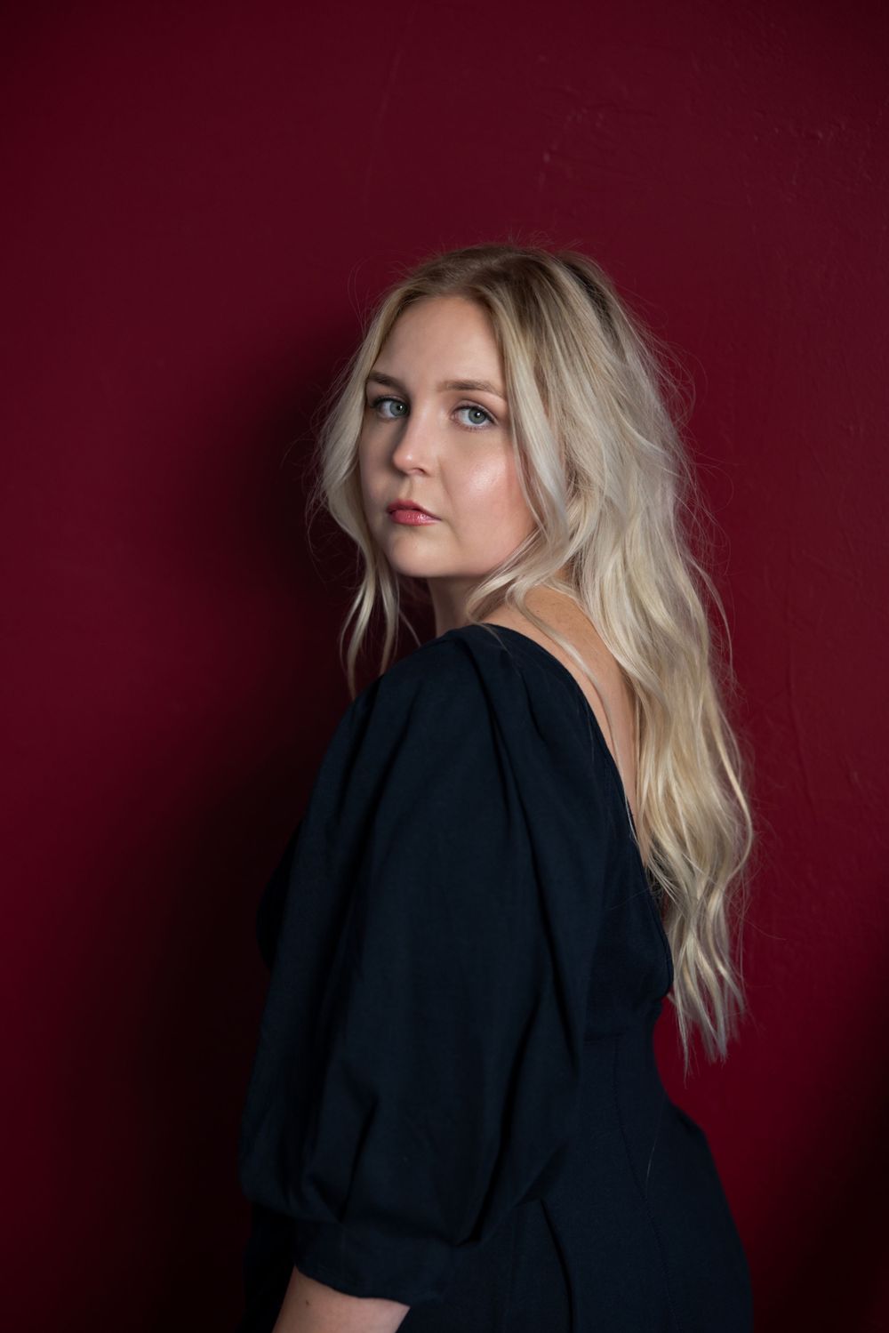 Portrait of a person with blonde wavy hair wearing a black top posing against a deep burgundy background.