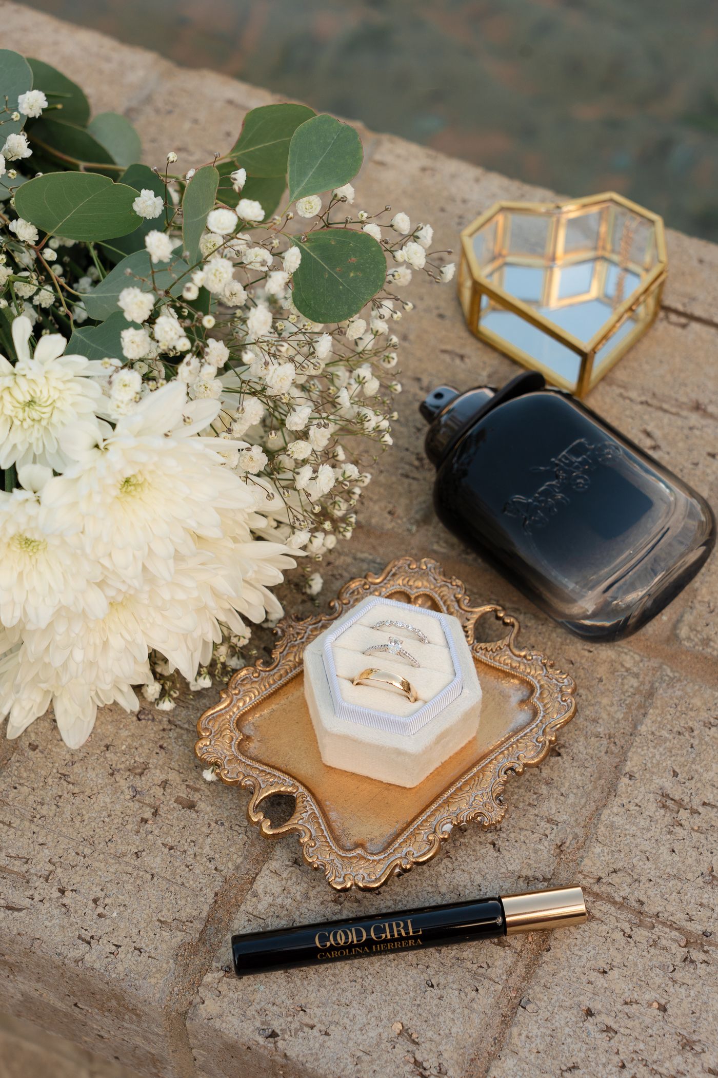 Wedding details including white flowers, ring box, and makeup items arranged on rustic surface.