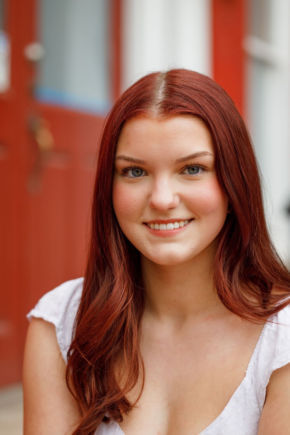 Portrait against orange wall featuring bright blue eyes and auburn hair wearing a white top.