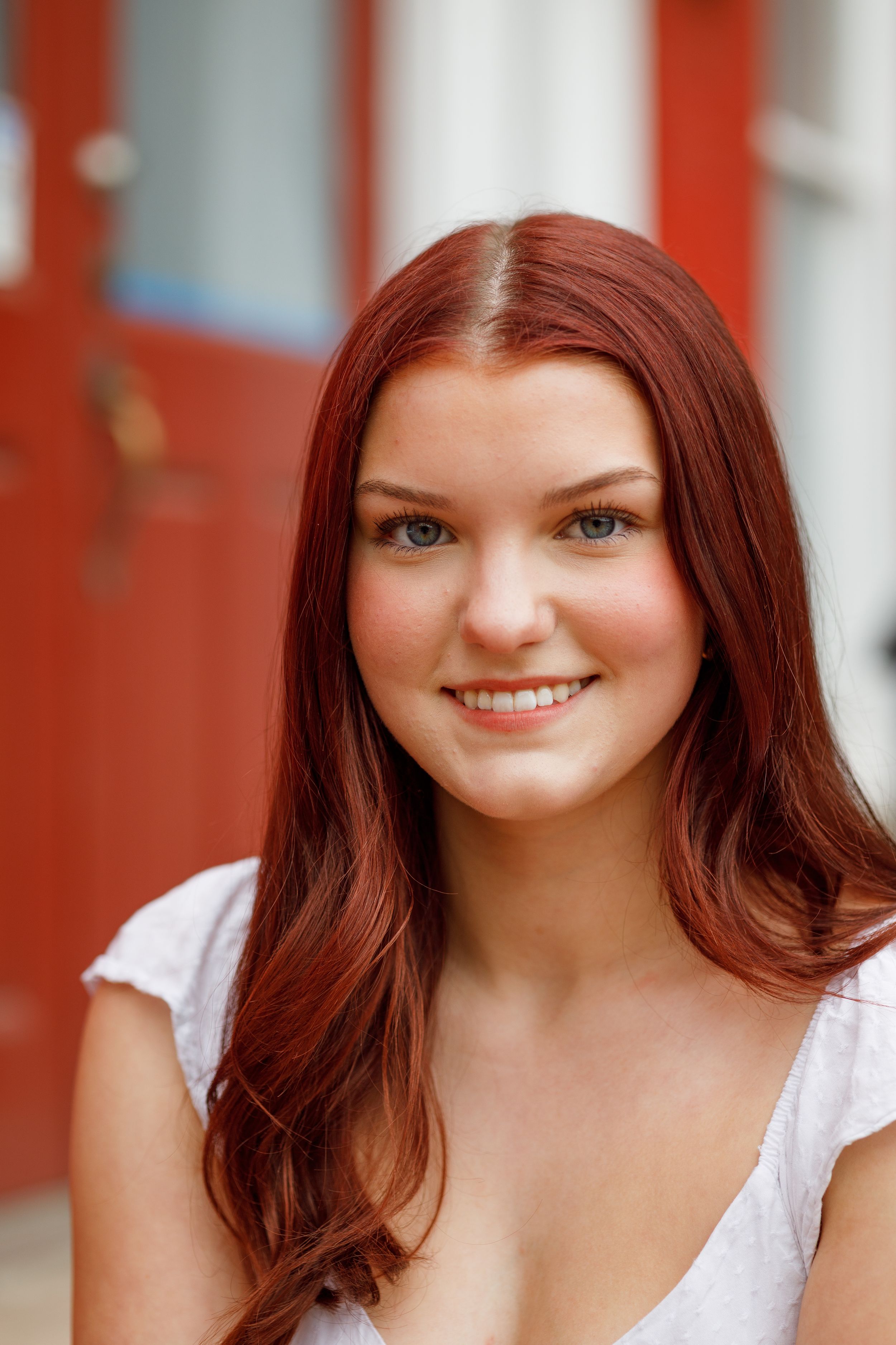 Portrait against orange wall featuring bright blue eyes and auburn hair wearing a white top.
