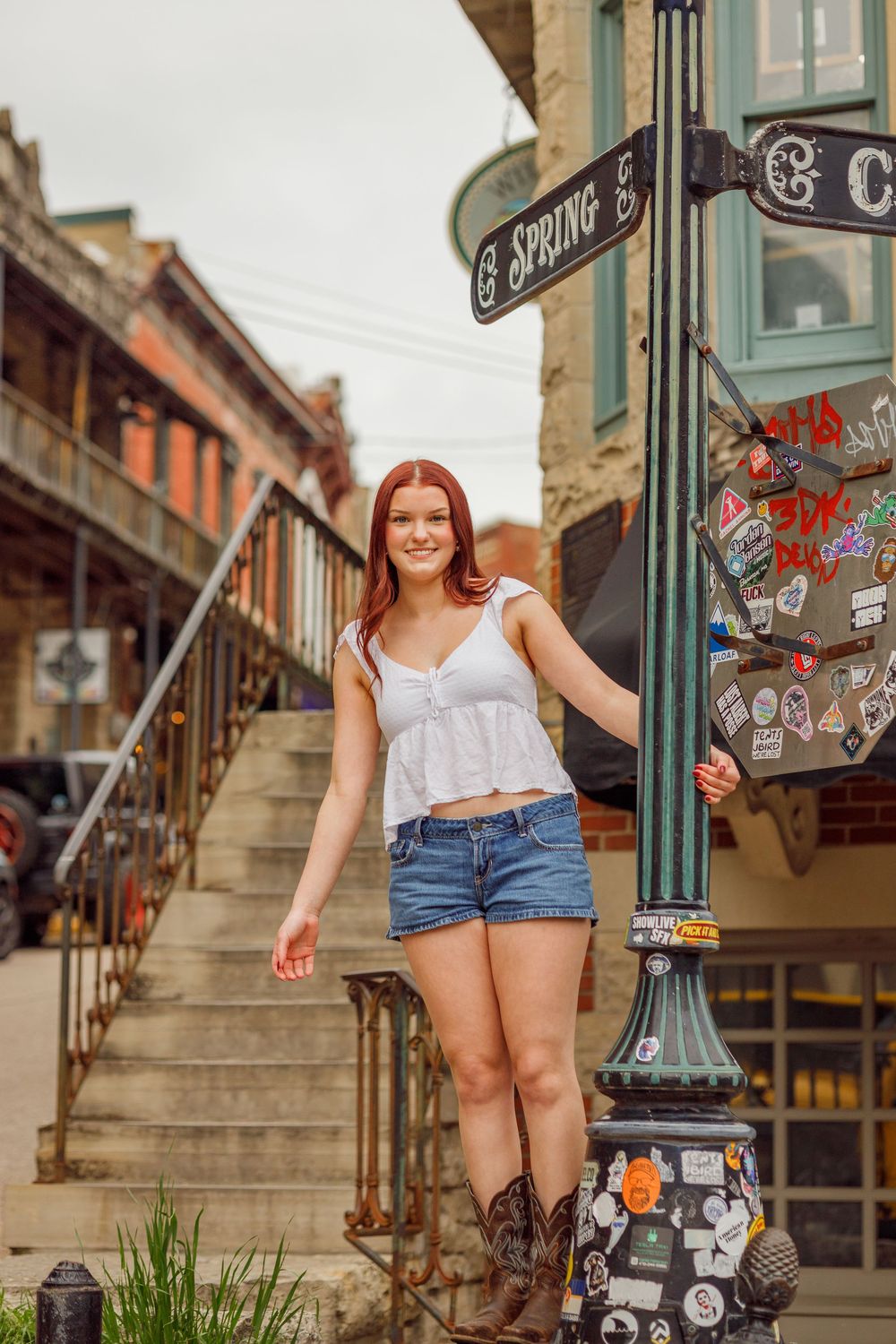 A woman wearing shorts and a white top sits casually on steps in a historic urban area beside a decorative lamp post.