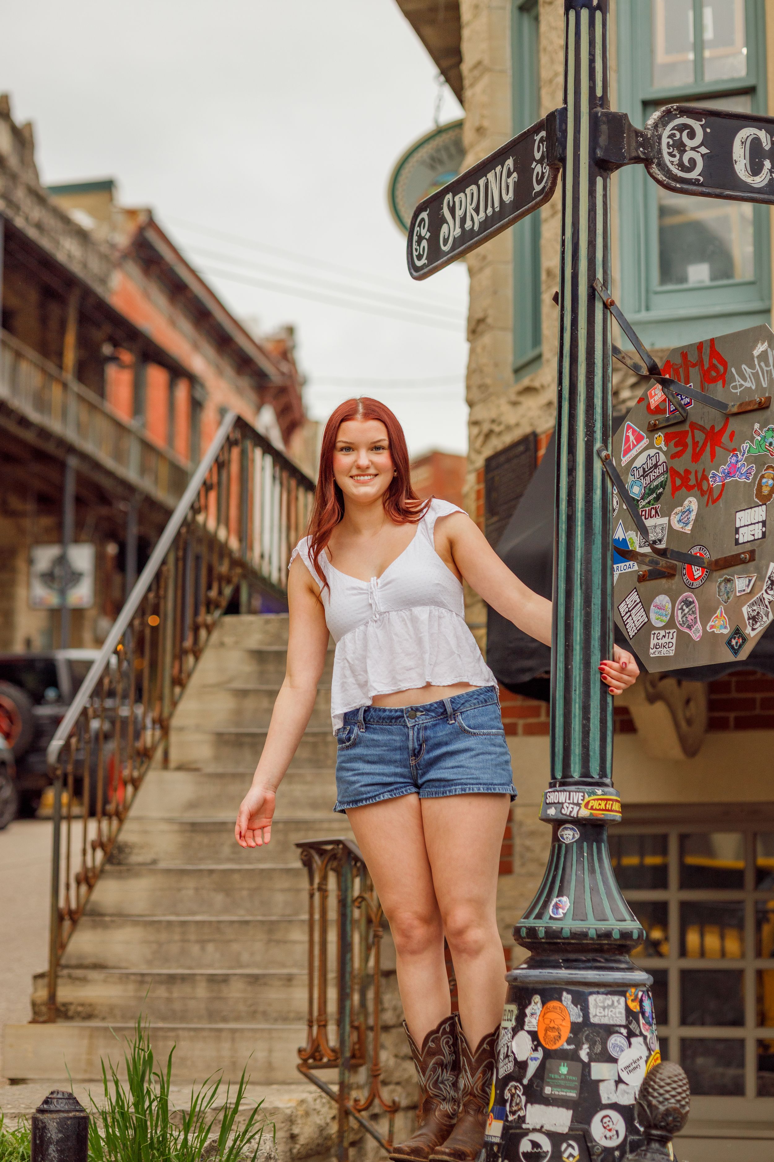 A woman wearing shorts and a white top sits casually on steps in a historic urban area beside a decorative lamp post.