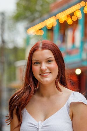 A portrait outdoors with white top and red hair against a background with warm bokeh lights and turquoise details.