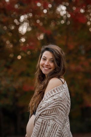 Woman in knit sweater smiles over her shoulder with autumn foliage in background.