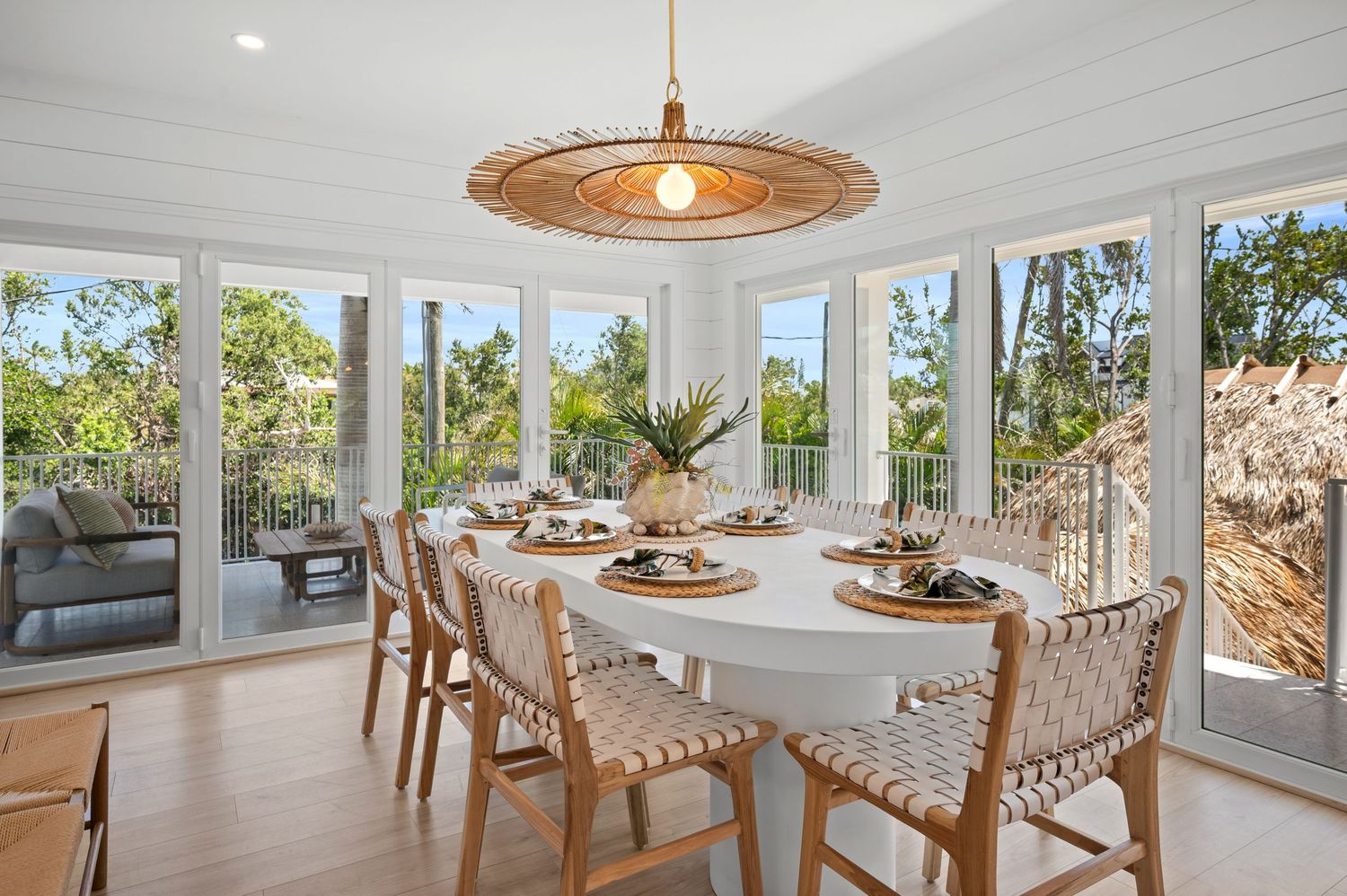 Modern coastal dining room with white table, woven chairs, and rattan pendant light overlooking tropical landscape through windows.