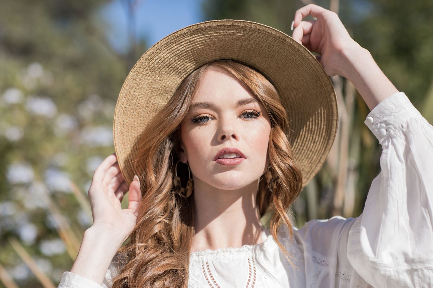 Young woman in a straw hat and white blouse poses outdoors in a sunny garden setting.