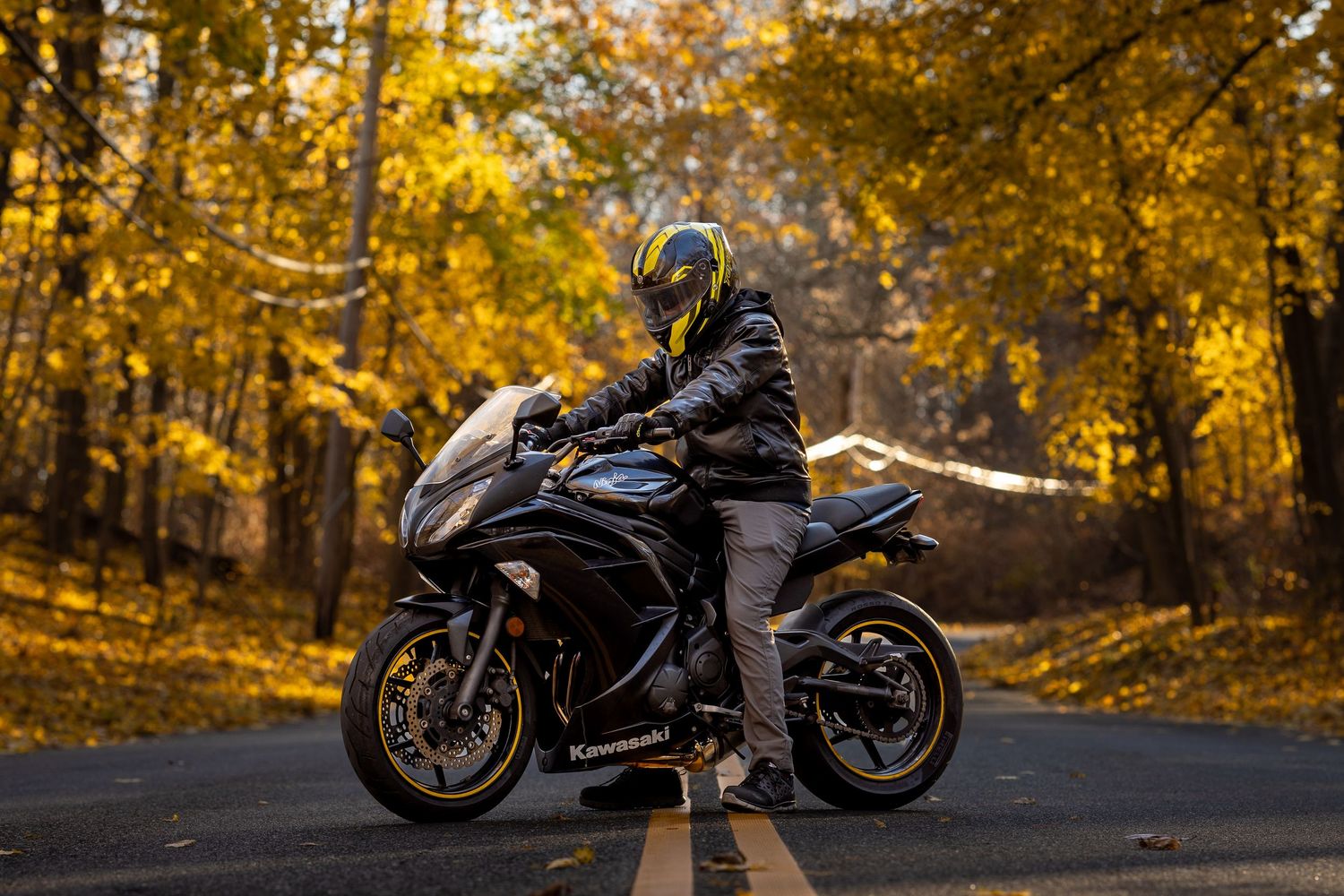 Person in leather jacket standing beside black sport motorcycle on coastal road under blue sky.