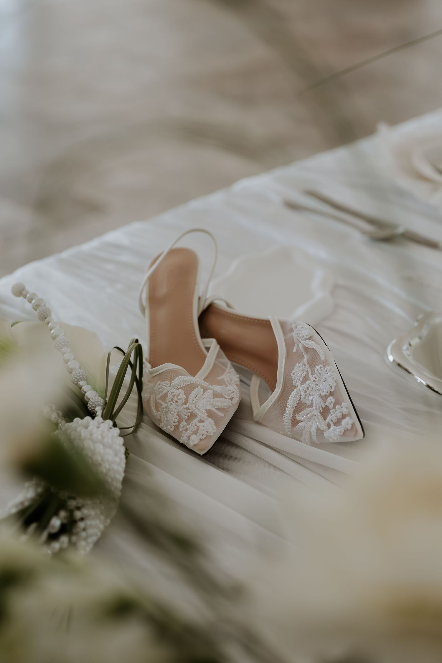 Elegant white lace bridal shoes with floral details lying on white fabric next to delicate flowers.