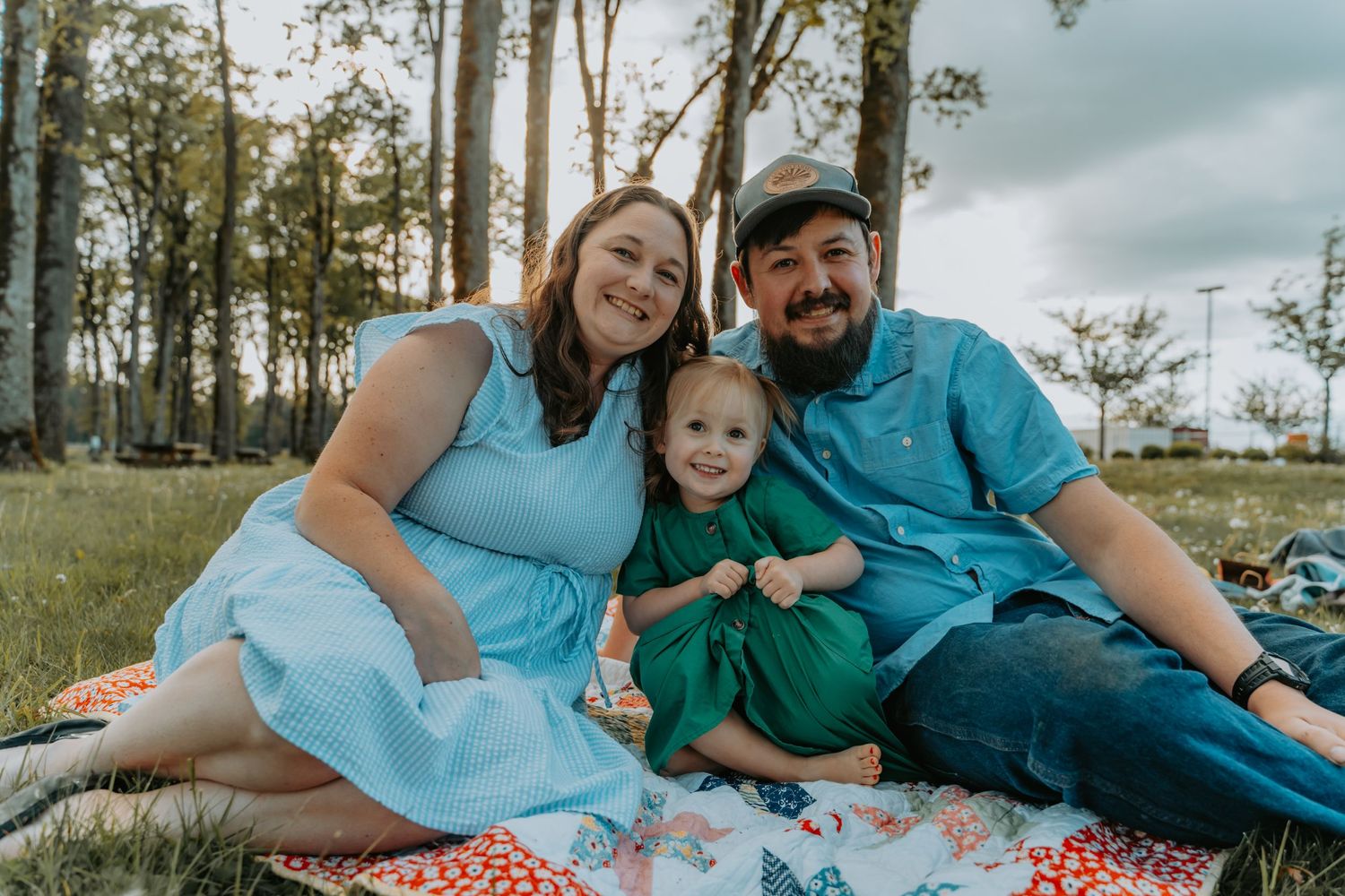 Family having a picnic on a blanket outdoors in a grassy area with trees in the background.