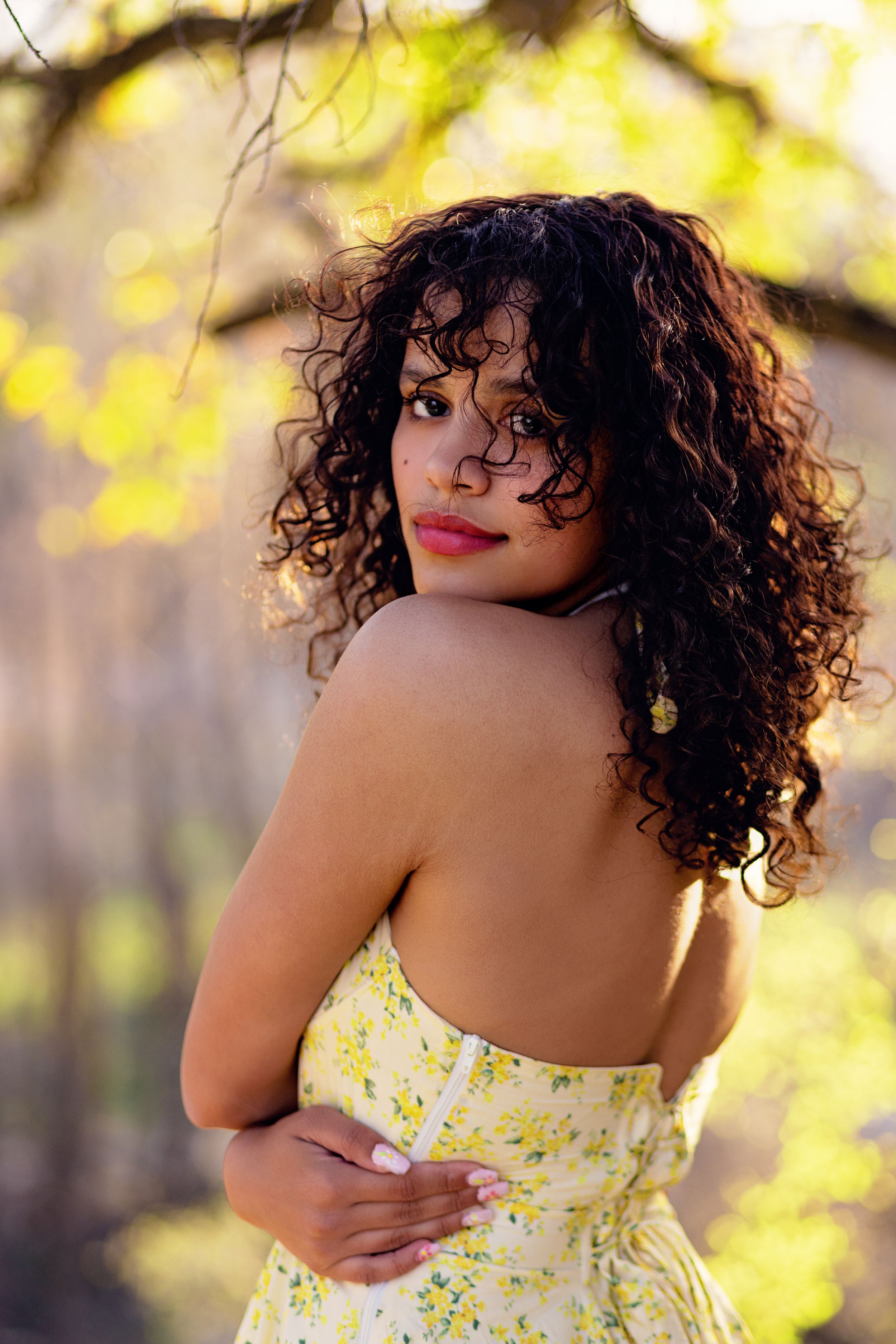 An elegant portrait in soft sunlight showing curly dark hair and a yellow floral dress against a dreamy bokeh background.
