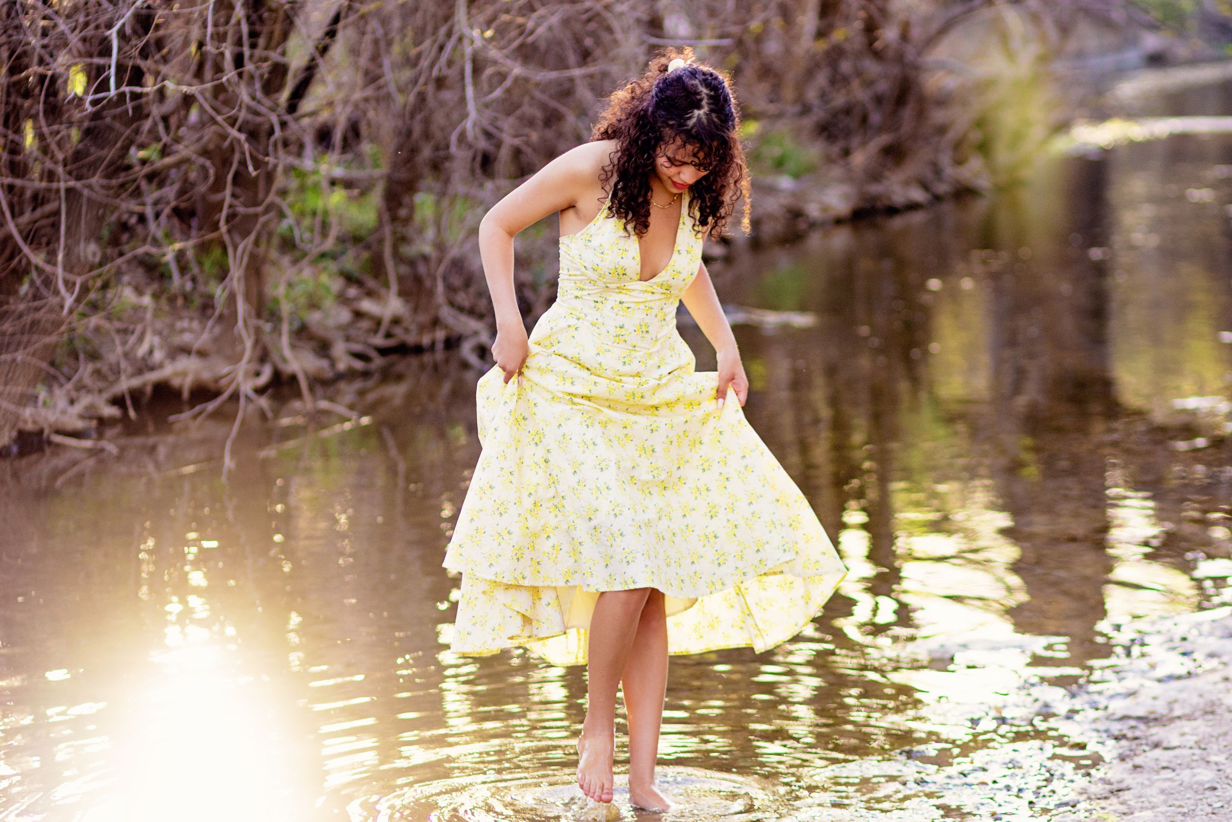 A person in a flowing yellow sundress stands playfully in shallow water near a wooded creek bank on a sunny day.