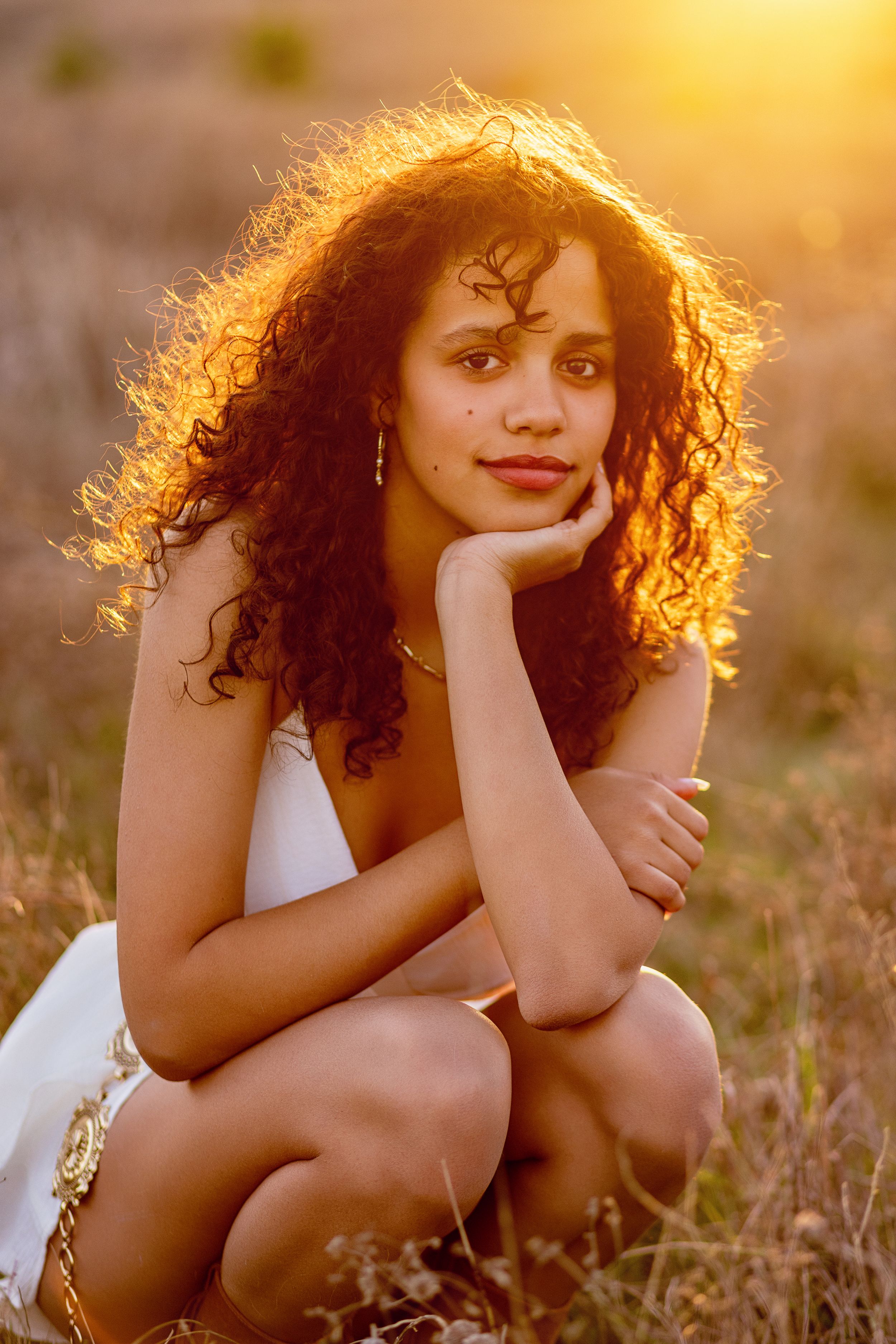 A woman with curly dark hair poses in a white dress during golden hour in a meadow at sunset.