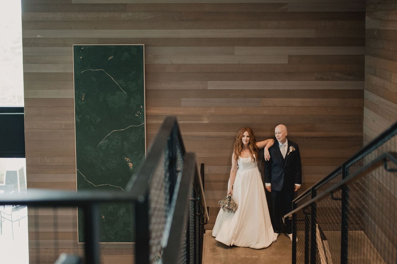 A formal couple descends a modern staircase with wooden walls and black railings.