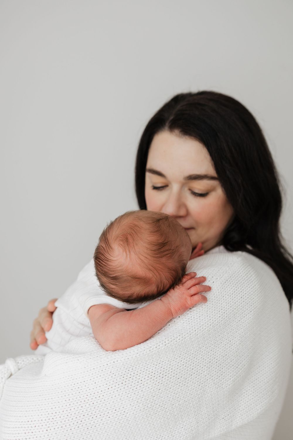 A mother with dark hair gently holds and kisses her newborn baby, both dressed in white.