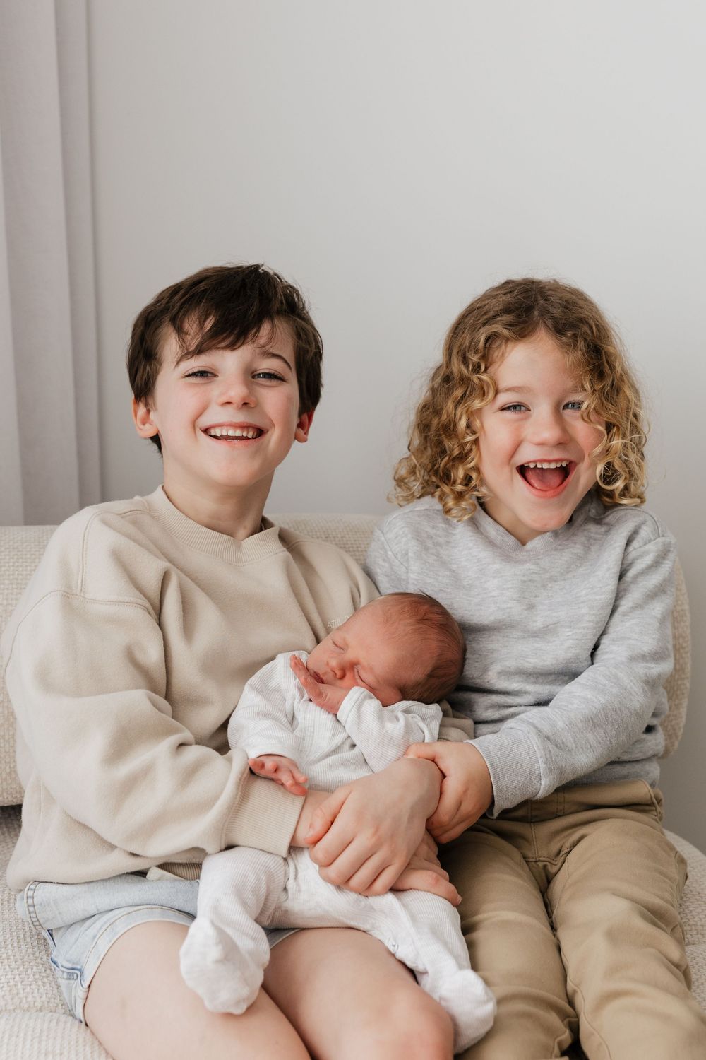Two smiling children sit on a couch holding a sleeping newborn baby sibling, sharing a joyful family moment.