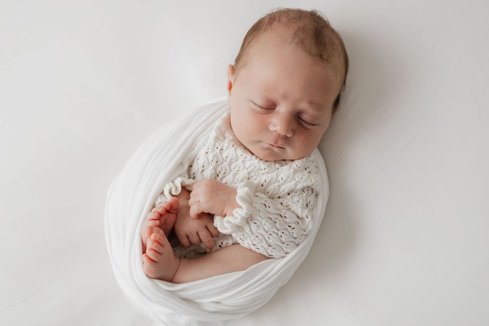 Peaceful newborn baby swaddled in white knit wrap sleeping on white background in professional photo.