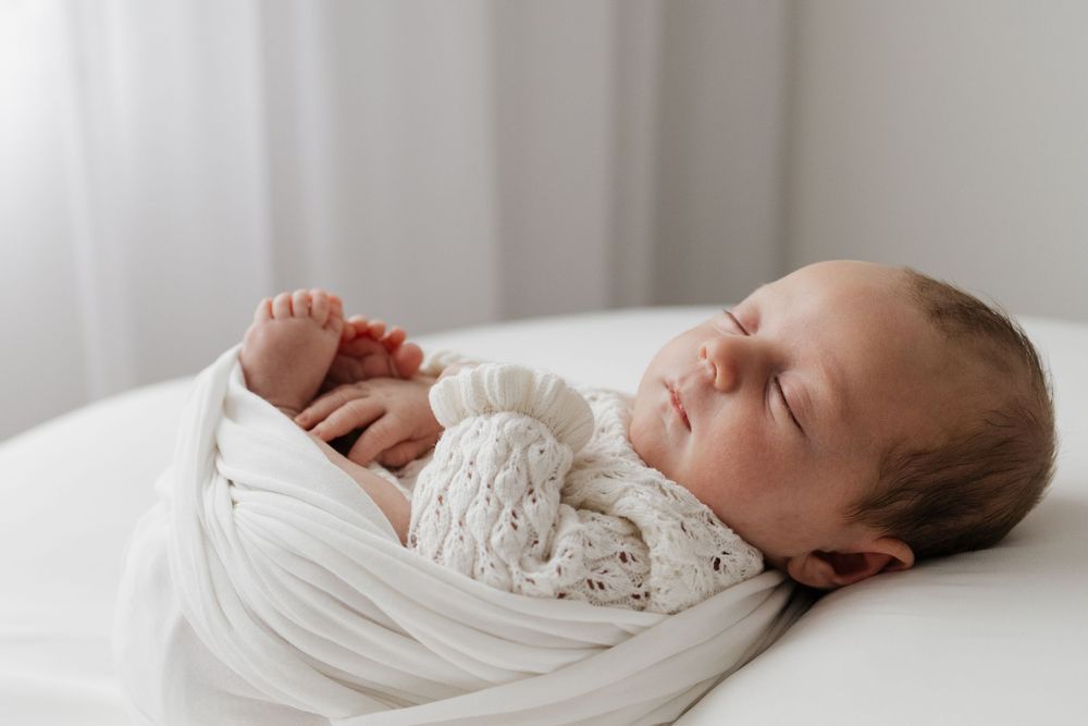Peaceful newborn baby sleeping swaddled in a soft white knit wrap on a white surface in a bright, airy room.