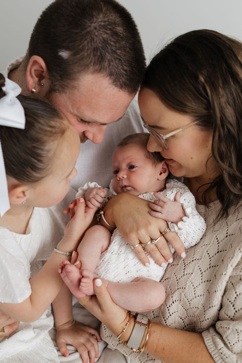 A family of four admires their newborn baby, with parents and young daughter in white clothing in a bright studio setting.