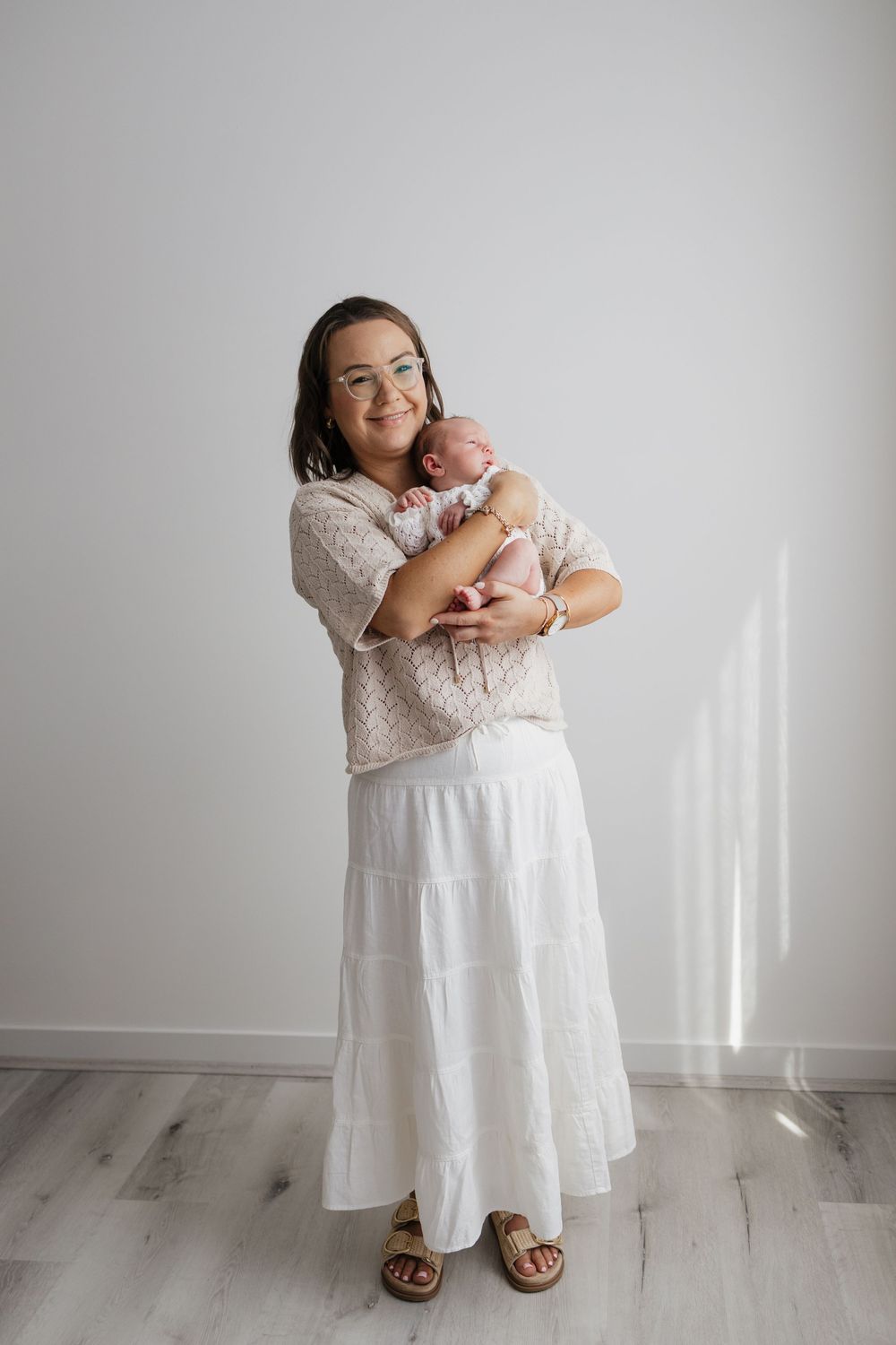 A smiling woman in a white skirt and floral top holds a newborn baby against a light background.