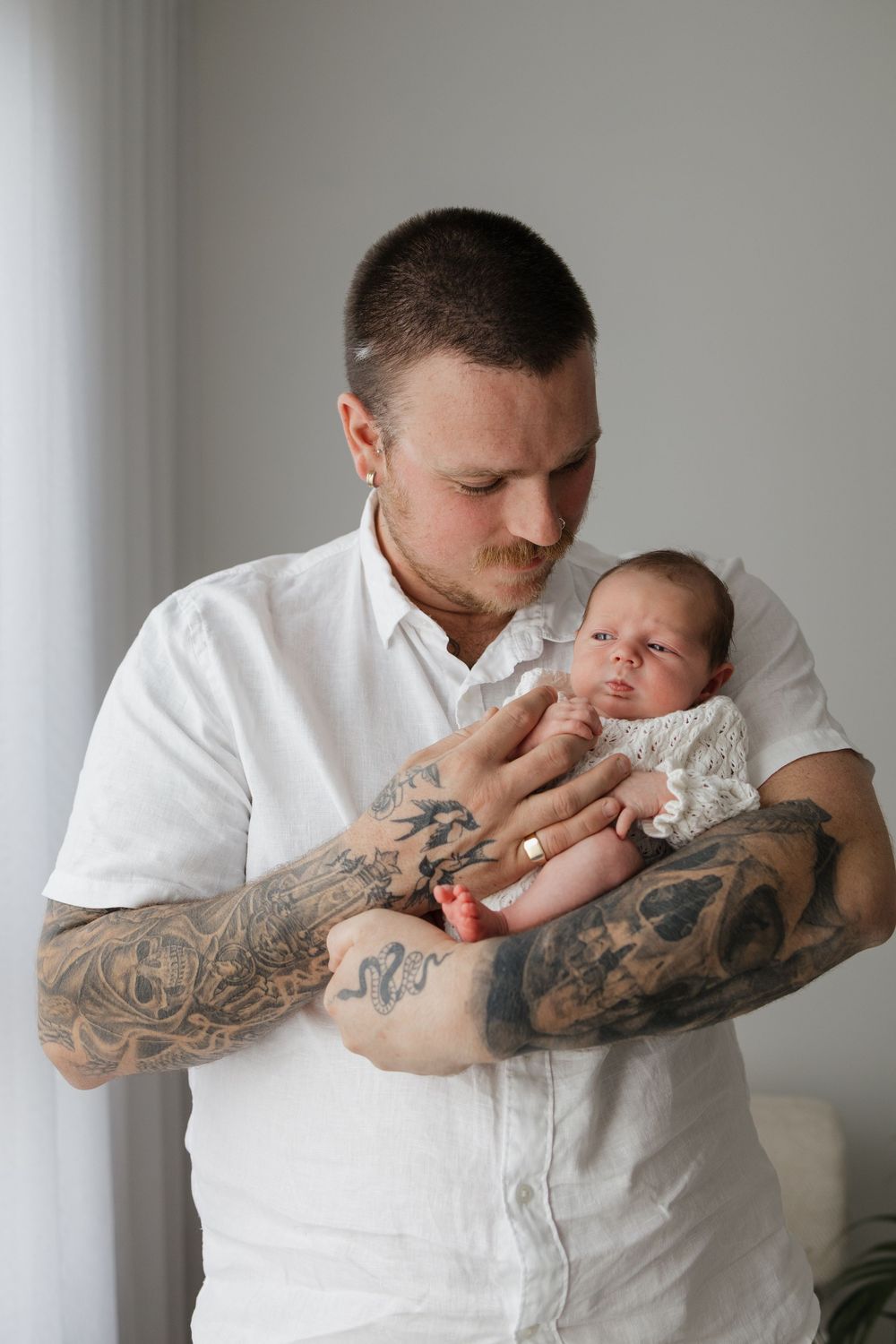 Tattooed father in white shirt gently holding his newborn baby, sharing a tender moment indoors.