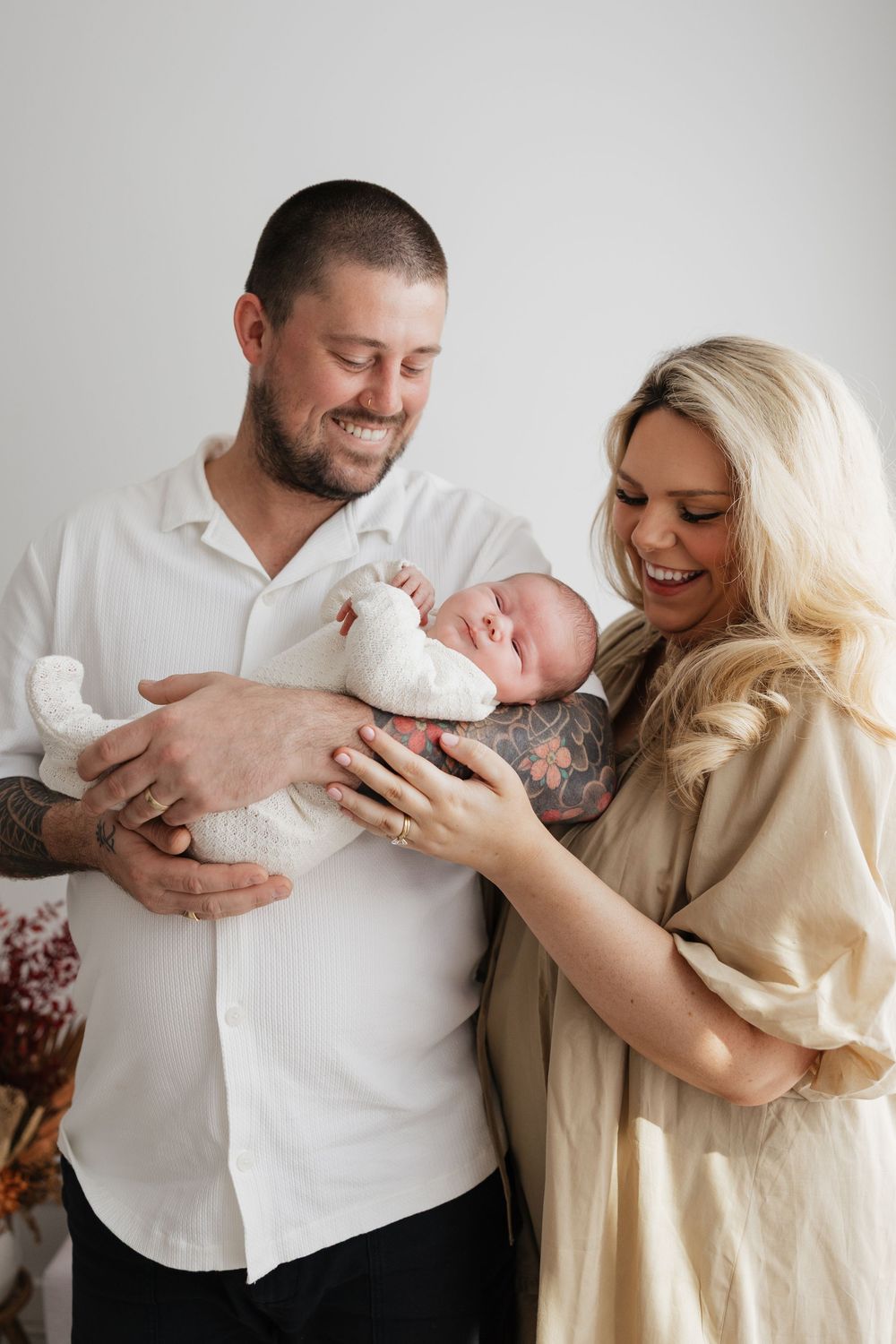 Smiling couple with tattoos holding their newborn baby wrapped in white, posing for a family portrait.