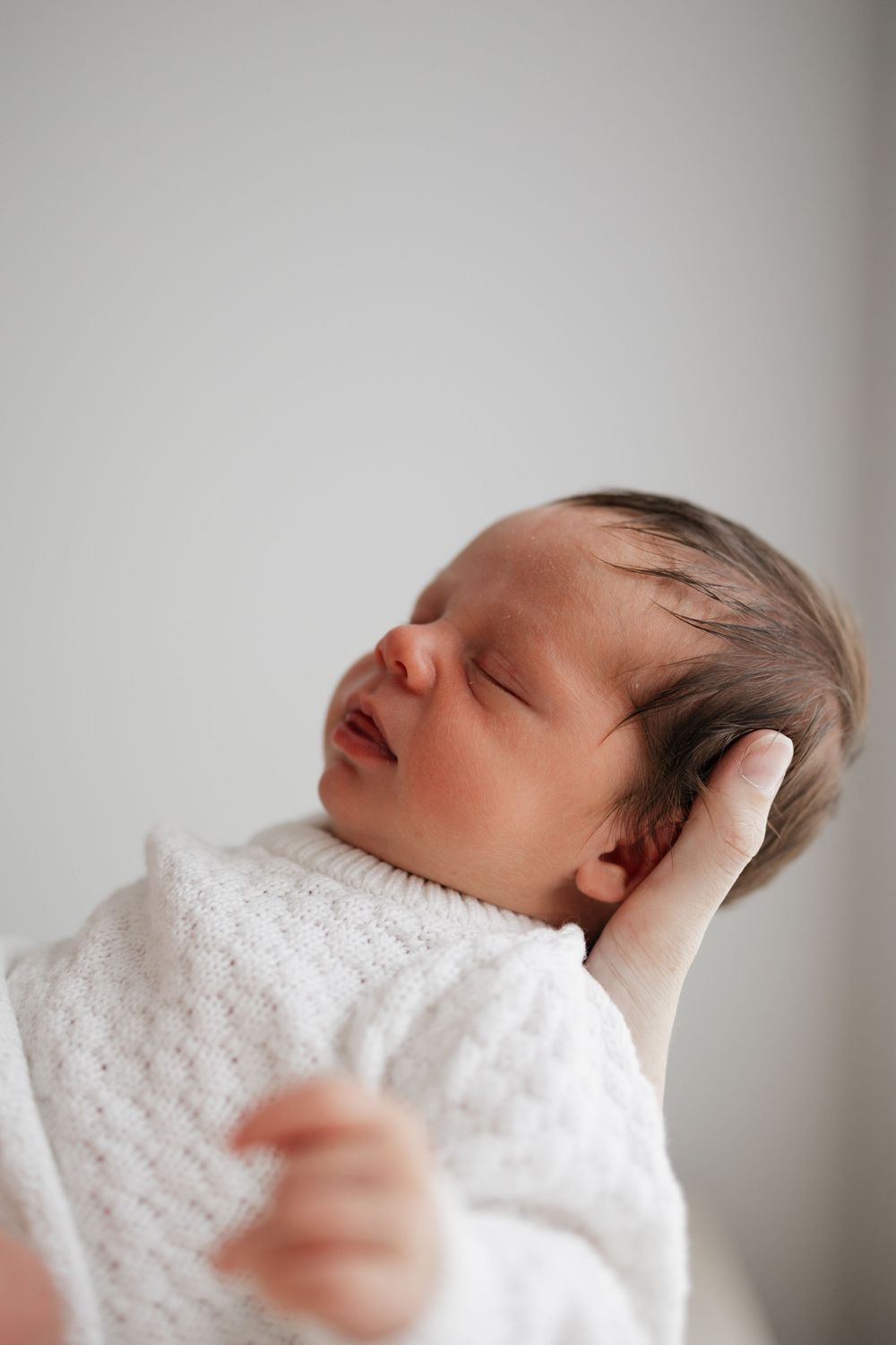 Peaceful newborn baby sleeping wrapped in a soft white knit blanket, with dark hair and tiny fingers visible.