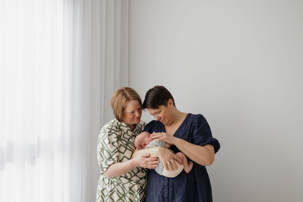 Two women admiring a sleeping newborn baby near a bright window in a soft, minimalist setting.