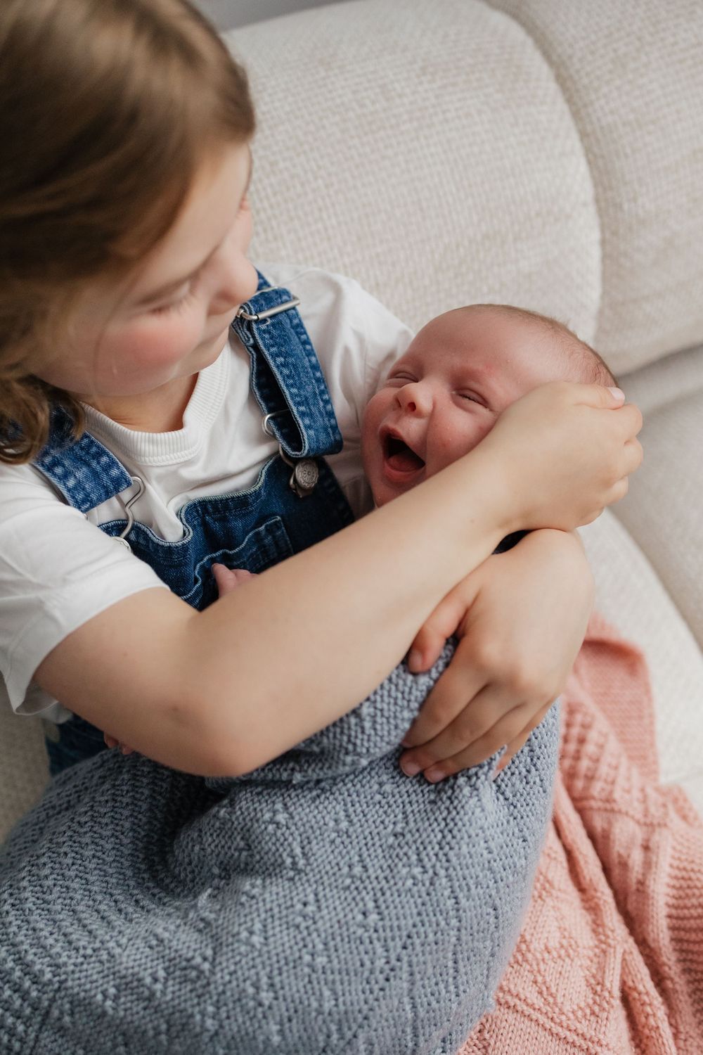 Young girl in denim overalls holding a yawning newborn baby wrapped in a knit blanket on a sofa.