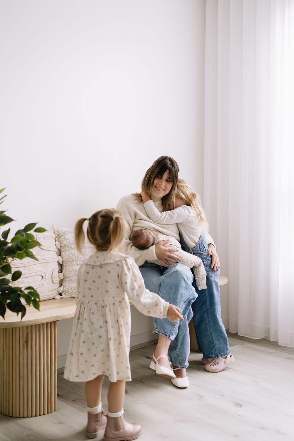 A mother sits on a bench holding a newborn while a toddler girl approaches in a bright, minimalist white room.