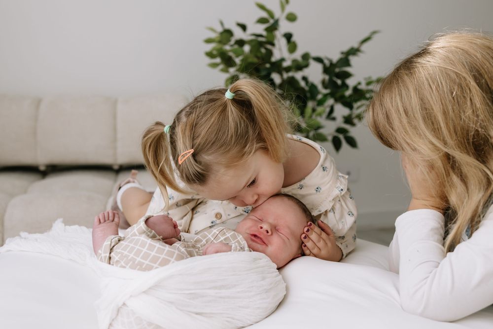 A young girl gently kisses a sleeping newborn baby while their mother watches lovingly on a white bed.