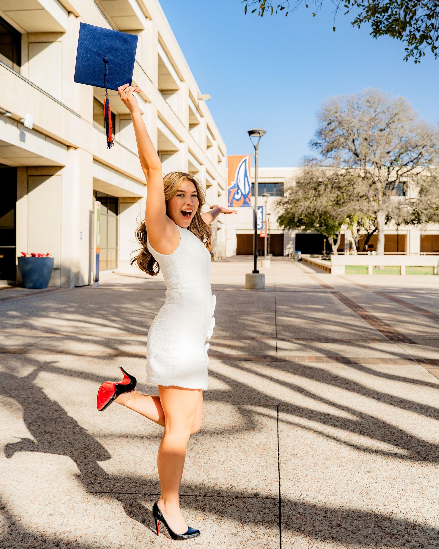 A graduate in white dress celebrates with excitement outside a university building.
