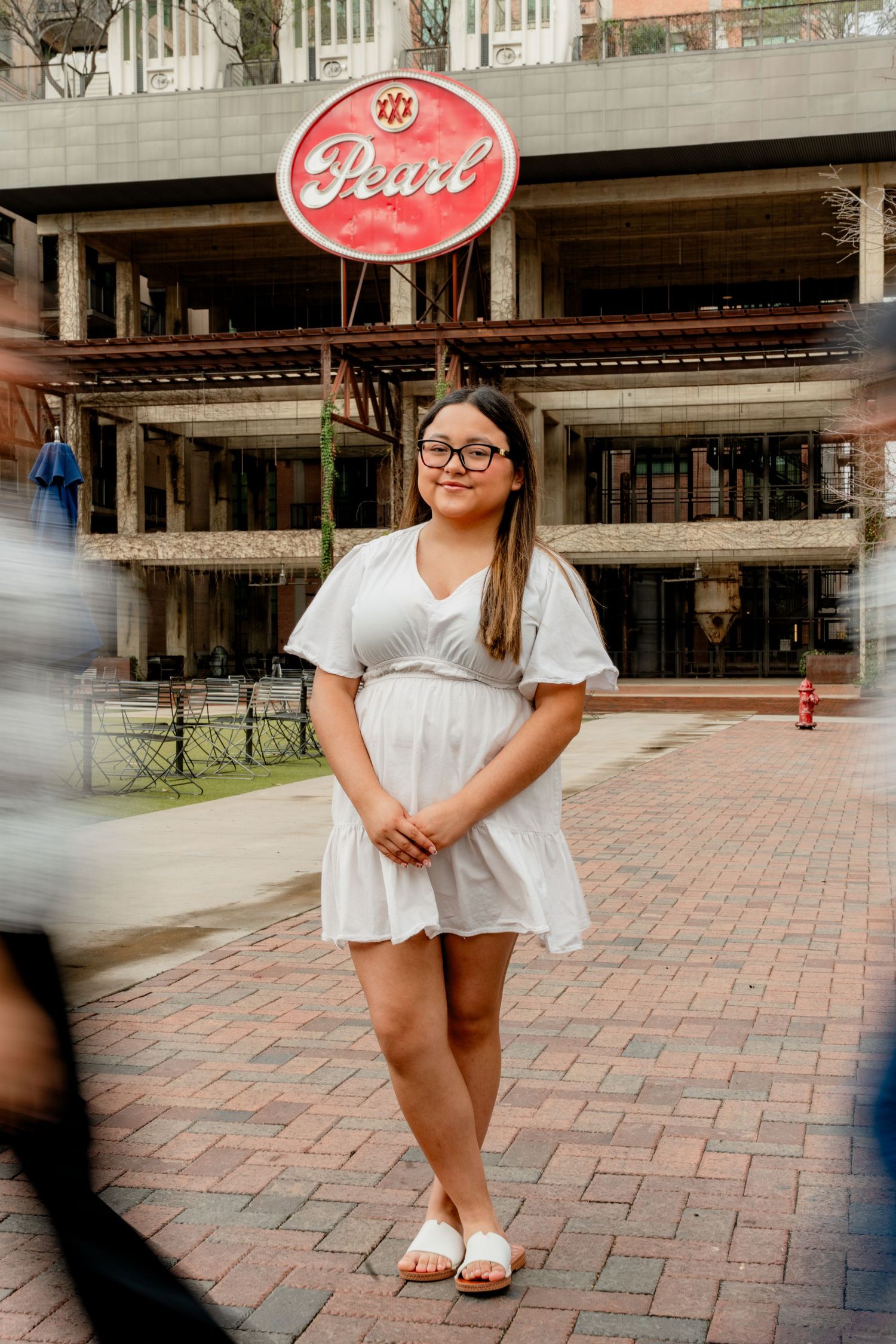 A person wearing a white dress and glasses stands on a brick walkway beneath a retro Pearl sign.